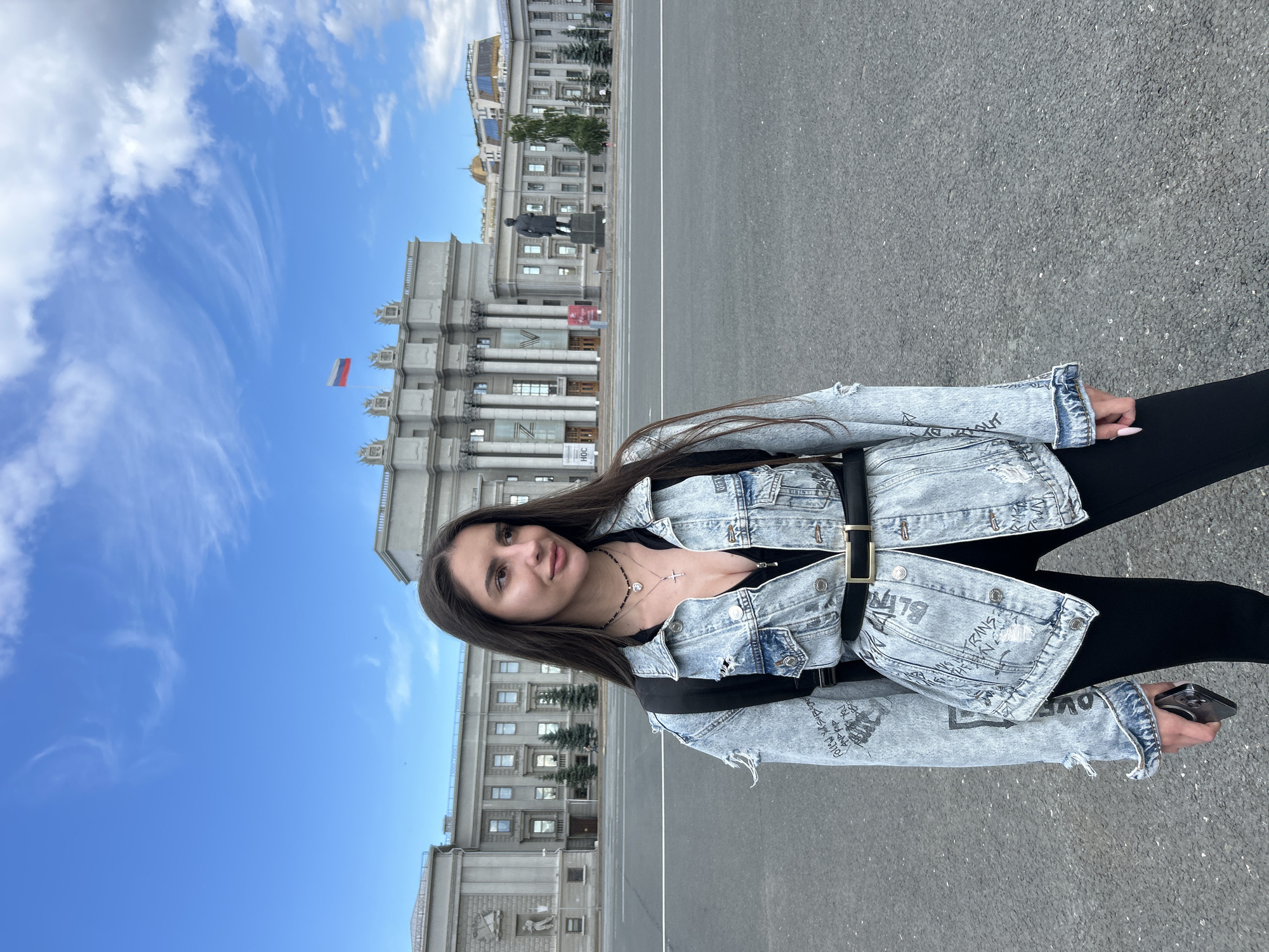 A young woman with long, dark hair poses for a photo in front of a grand, neoclassical building under a bright blue sky with wispy clouds. She is wearing a light-wash denim jacket with distressed details and black lettering, a black top, and black pants. A black belt with a gold buckle cinches her waist, and she has a delicate silver necklace with a cross pendant. In her right hand, she holds a smartphone.
The building behind her is large and imposing, with multiple floors, columns, and ornate architectural details. A Russian flag is visible flying from a flagpole on the building's facade. A statue of a man on a pedestal stands in the foreground, slightly to the left of the center. The ground in front of the building is a paved surface, likely a plaza or street. The scene appears to be set during the daytime, under clear weather. The overall mood of the image is casual and confident. Some visible text on the woman's jacket appears to be in English, with words like "BLING" and "LOVE" partially discernible. The location is identified as Novokuybyshevsk, Russia.