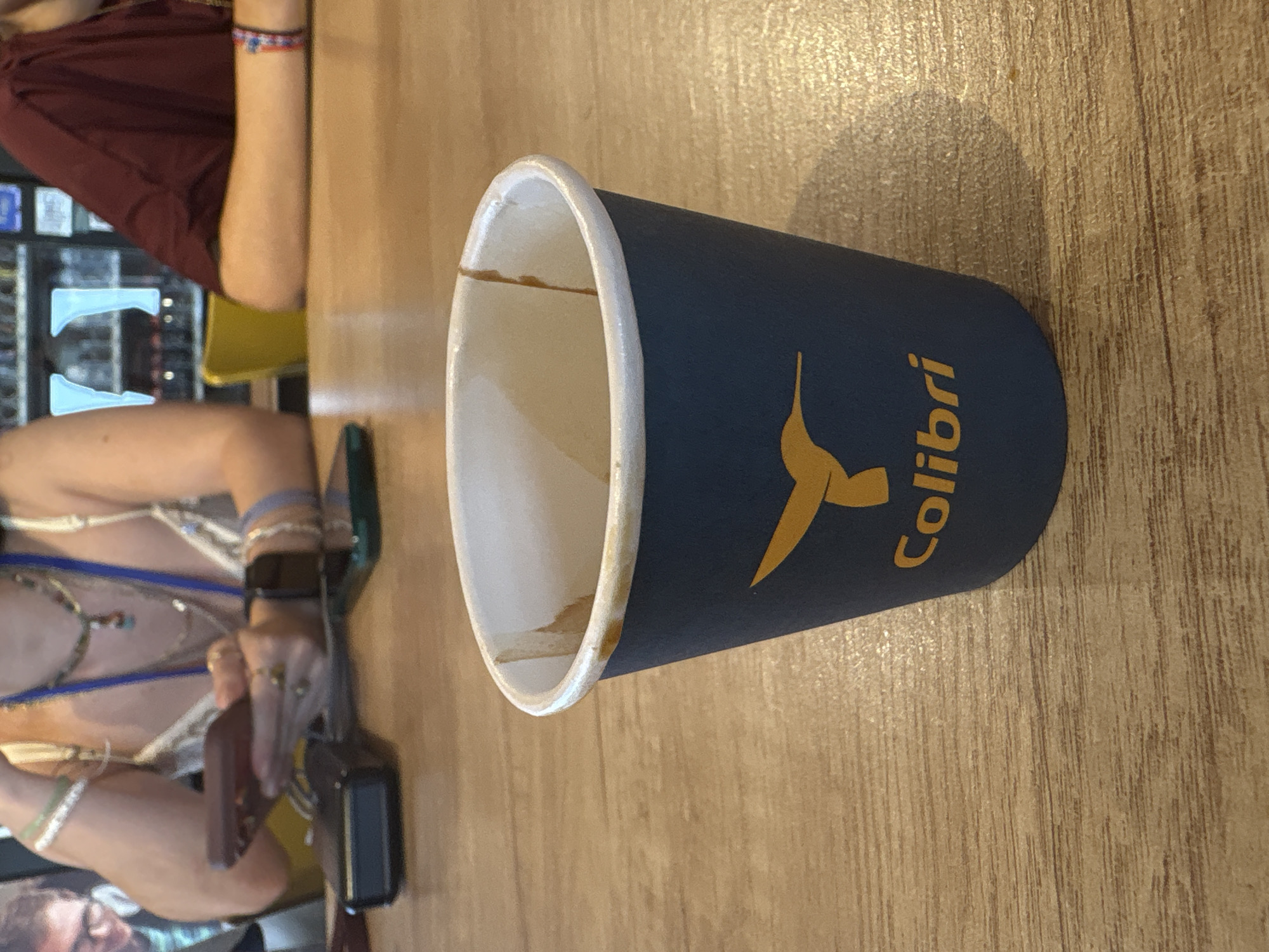 The image depicts a close-up view of a dark blue paper cup with a yellow hummingbird logo and the word "Colibri" printed on it. The cup is placed on a light brown wooden table. The inside of the cup shows remnants of a brown liquid, possibly coffee or tea, with some dried drips along the rim. In the background, out of focus, are two individuals. One person, on the left, is wearing jewelry and holding a smartphone. Another person, on the right, is wearing a dark red shirt. The overall setting appears to be an indoor seating area, possibly a cafe or restaurant, with a blurry background suggesting a beverage cooler. The image does not contain any specific visual cues that would pinpoint its location to Almodôvar, Portugal.