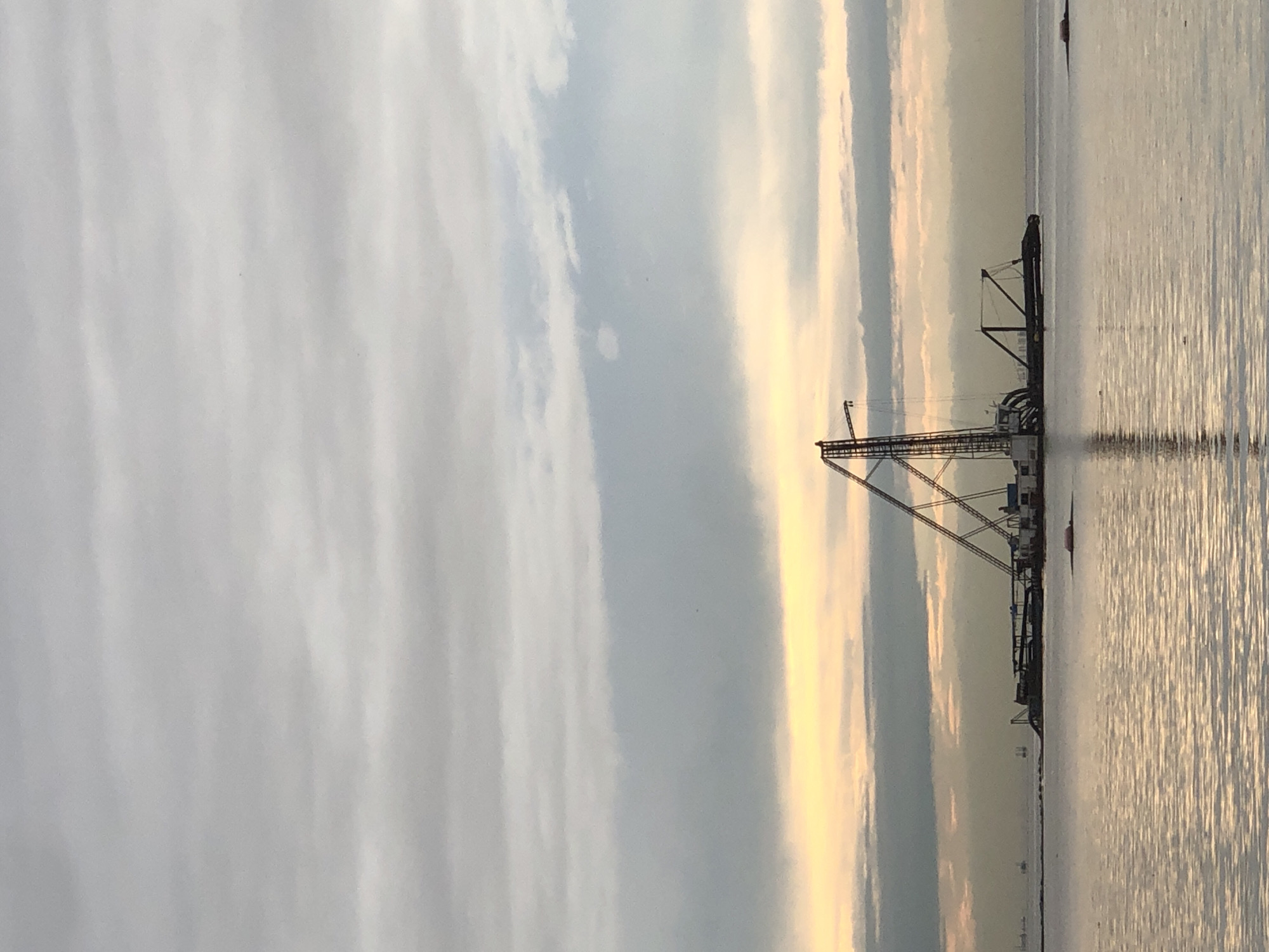 A wide-angle shot captures a hazy, overcast sky over a body of water, possibly the Lagos Lagoon near Ado, Nigeria. In the middle distance, a large dredging vessel is moored, its cranes and equipment silhouetted against the muted light. The water reflects the sky, creating shimmering patterns. In the far distance, several smaller vessels and oil rigs are visible on the horizon. The overall atmosphere is somber and industrial, with a hint of dawn or dusk lighting the scene.