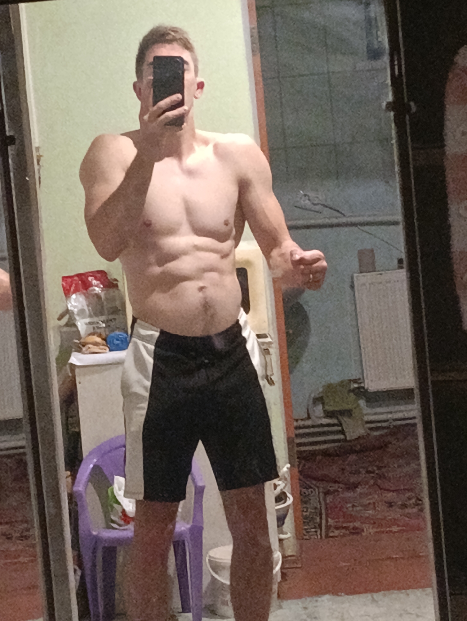 A man is taking a mirror selfie in a dimly lit room. He is shirtless, wearing black and white athletic shorts. His physique is muscular, with visible abs and chest muscles. He is holding a smartphone in front of his face, obscuring it from view. The room appears to be a bedroom or a personal space, with a bed, a radiator, and a chair visible in the reflection. The walls are a pale green color. In the foreground, a purple plastic chair is visible with some items on it. The overall impression is that of a casual, personal moment captured by the individual himself.