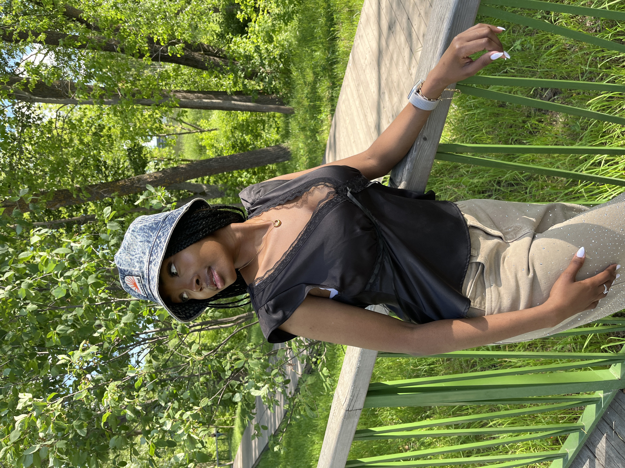 A young Black woman wearing a denim bucket hat, a black lace-trimmed V-neck top, and beige pants adorned with rhinestones, stands leaning on a green-railed wooden bridge in a park. She has long braided black hair and is looking to her left. The background features lush green trees and a wooden boardwalk. The setting appears to be a public park with dense foliage, suggesting a natural or semi-natural environment within a city.