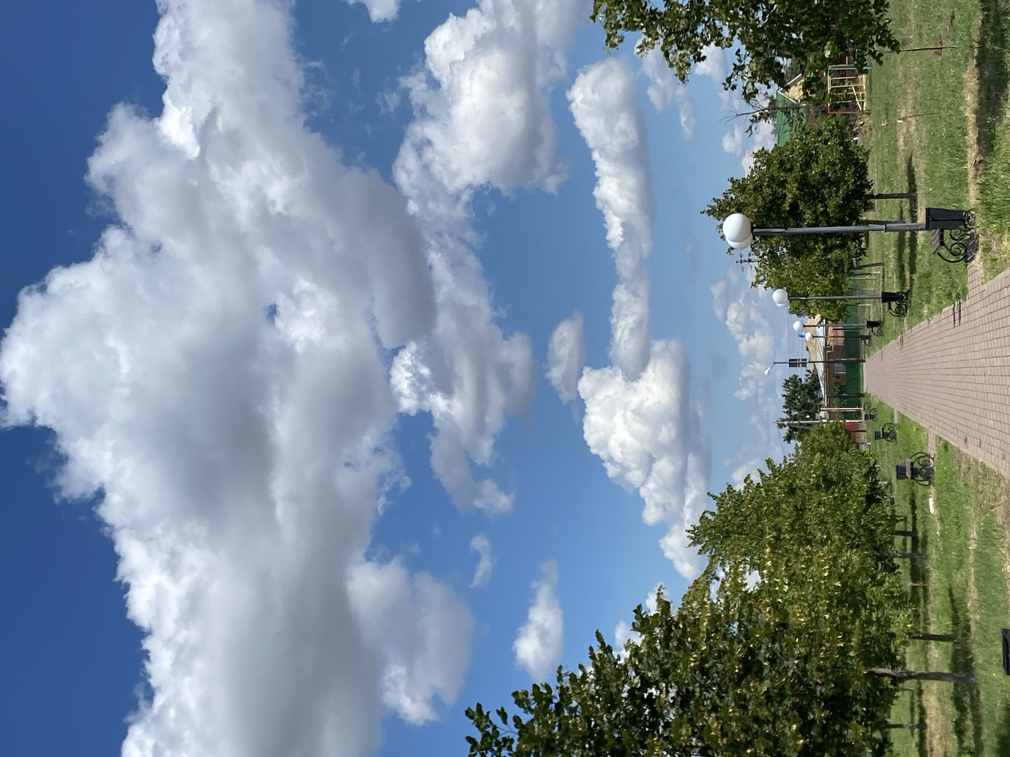 The image depicts a daytime scene in Kushchovskaya, Russia, featuring a park or public walkway. A brick-paved path leads into the distance, flanked on either side by mature trees with dense green foliage. Park benches and trash receptacles are visible along the path. The sky is a clear blue, dotted with large, fluffy white cumulus clouds. In the background, hints of buildings and what appears to be a playground structure can be seen. The lighting suggests it is a sunny day.