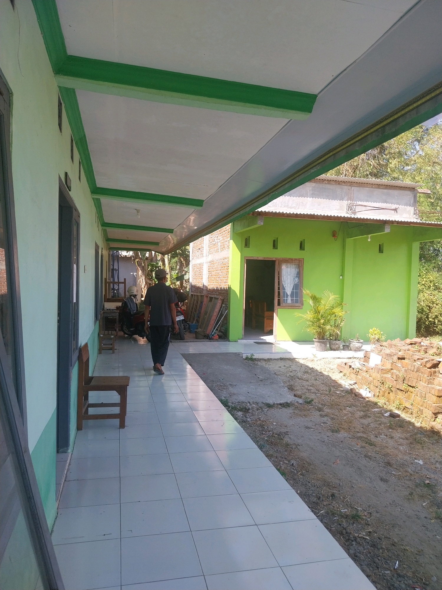 An exterior shot captures a sunlit scene in Pandean, Indonesia, focusing on a covered walkway adjacent to a bright green building. A lone male figure, wearing a dark t-shirt and black pants, walks away from the camera down the tiled walkway. To his left, a light green wall with dark-framed windows runs along the building. A simple wooden chair sits empty on the walkway. A red motorcycle is parked further down the walkway. The green building features an open doorway with a white curtained window. Potted plants, including a palm, are placed outside the entrance. To the right of the building, a pile of bricks and dry earth are visible. The scene is completed by a corrugated metal roof on the building and trees in the background.