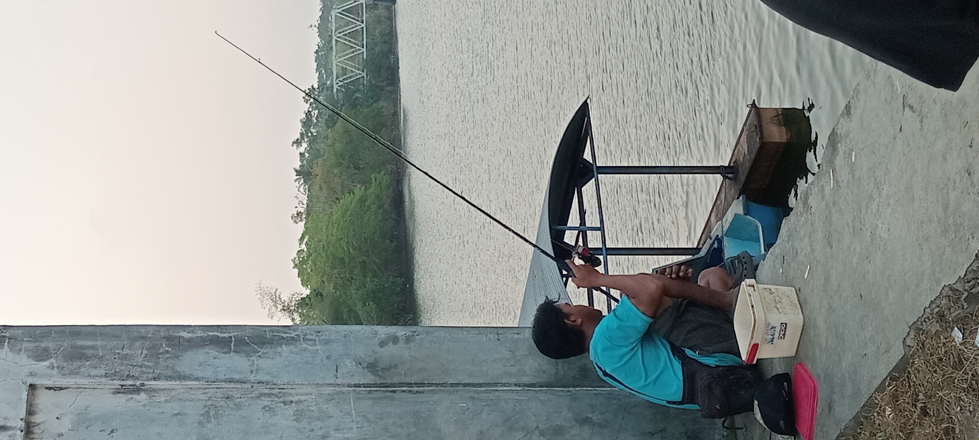 In Kalipucang, Indonesia, a person is seen fishing from a concrete embankment next to a body of water. The individual, wearing a blue t-shirt and dark shorts, sits with their legs tucked beneath them, holding a fishing rod. A dark bag is slung over their shoulder, and a white bucket with red trim is positioned in front of them. The water appears calm, with ripples visible. In the background, lush green trees line the opposite bank of the water, and a white bridge with a distinctive W-shaped structure is visible in the distance. The sky is overcast and light-colored. A portion of a large, weathered concrete pillar is prominent on the left side of the frame.