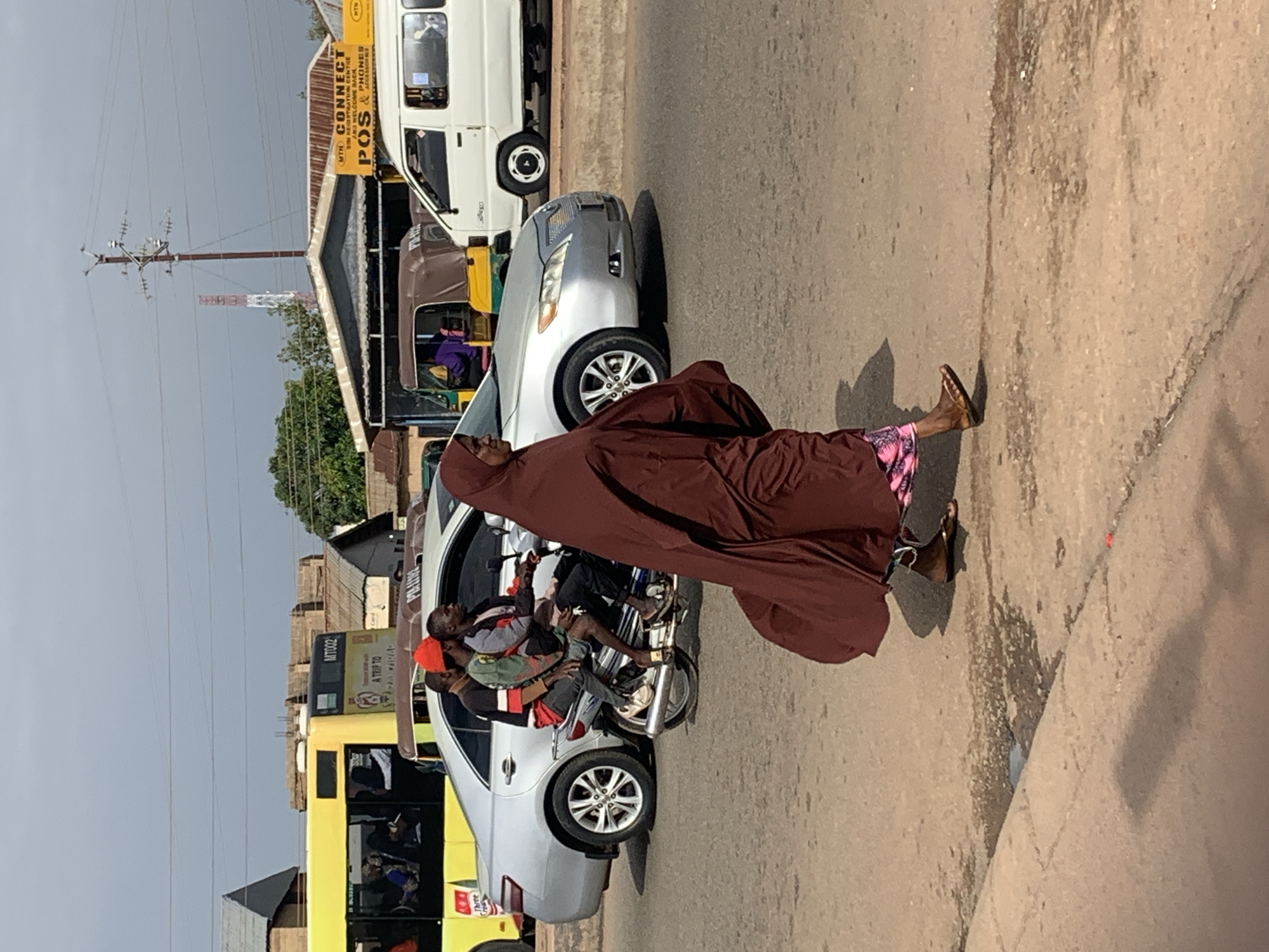 In Gura Top, Nigeria, a woman in a dark maroon hijab and a patterned skirt walks on a paved road. She is wearing brown sandals and a pink patterned fabric is visible beneath her skirt. Behind her, a silver sedan is parked on the road. A motorcycle with three male passengers, one wearing a bright orange cap, is also on the road, positioned between the sedan and the woman. Further back, a yellow bus is visible, and beyond that, several buildings and a white van are present. The sky is overcast.