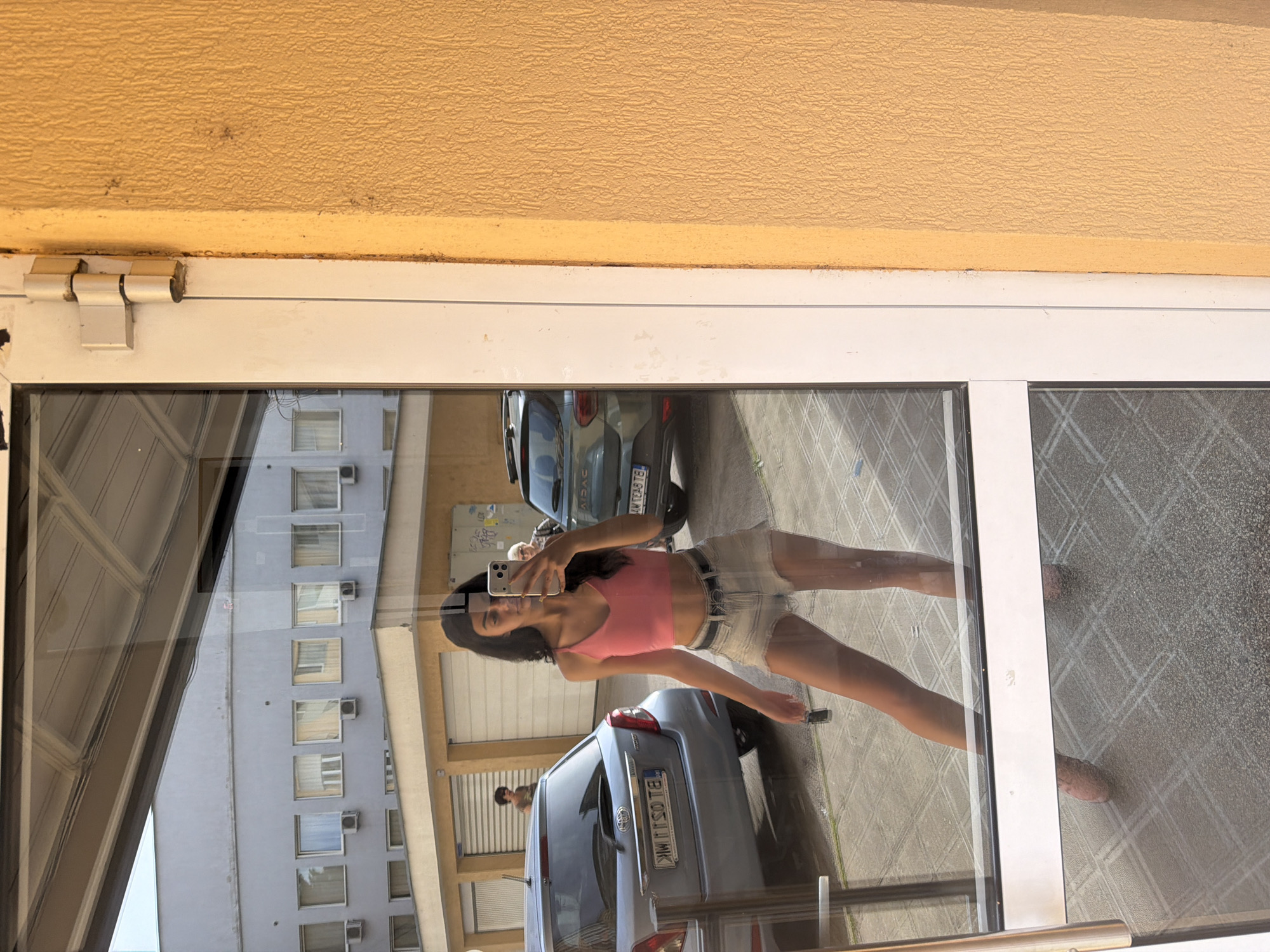 The image shows a young woman in a pink tank top and denim shorts taking a selfie in a reflection on a glass door. She is holding a phone in her right hand and a small object, possibly keys, in her left. She is wearing patterned pink shoes. Behind her, in the reflection, are two cars, one light blue and one dark green, and a building with multiple windows. The exterior of the building to the right of the door is a textured yellow-orange wall. The scene appears to be outdoors in an urban environment. The location is the city of Veliko Tarnovo, Bulgaria.
