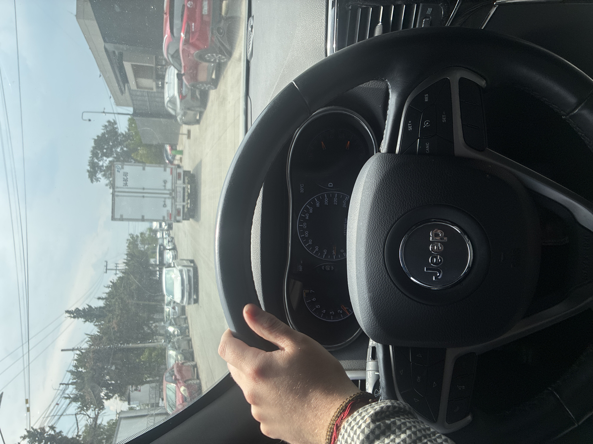 The image shows a person driving a Jeep, likely in Cuautitlán Izcalli, Mexico, based on the presence of Mexican license plates. The driver's hand is visible on the steering wheel. The dashboard and the road ahead with several cars and buildings are also visible. The steering wheel has the "Jeep" logo and cruise control buttons. The dashboard shows a temperature of 30°C. The car is in drive mode, indicated by "D" on the dashboard. There are electrical wires visible on the left side of the image.