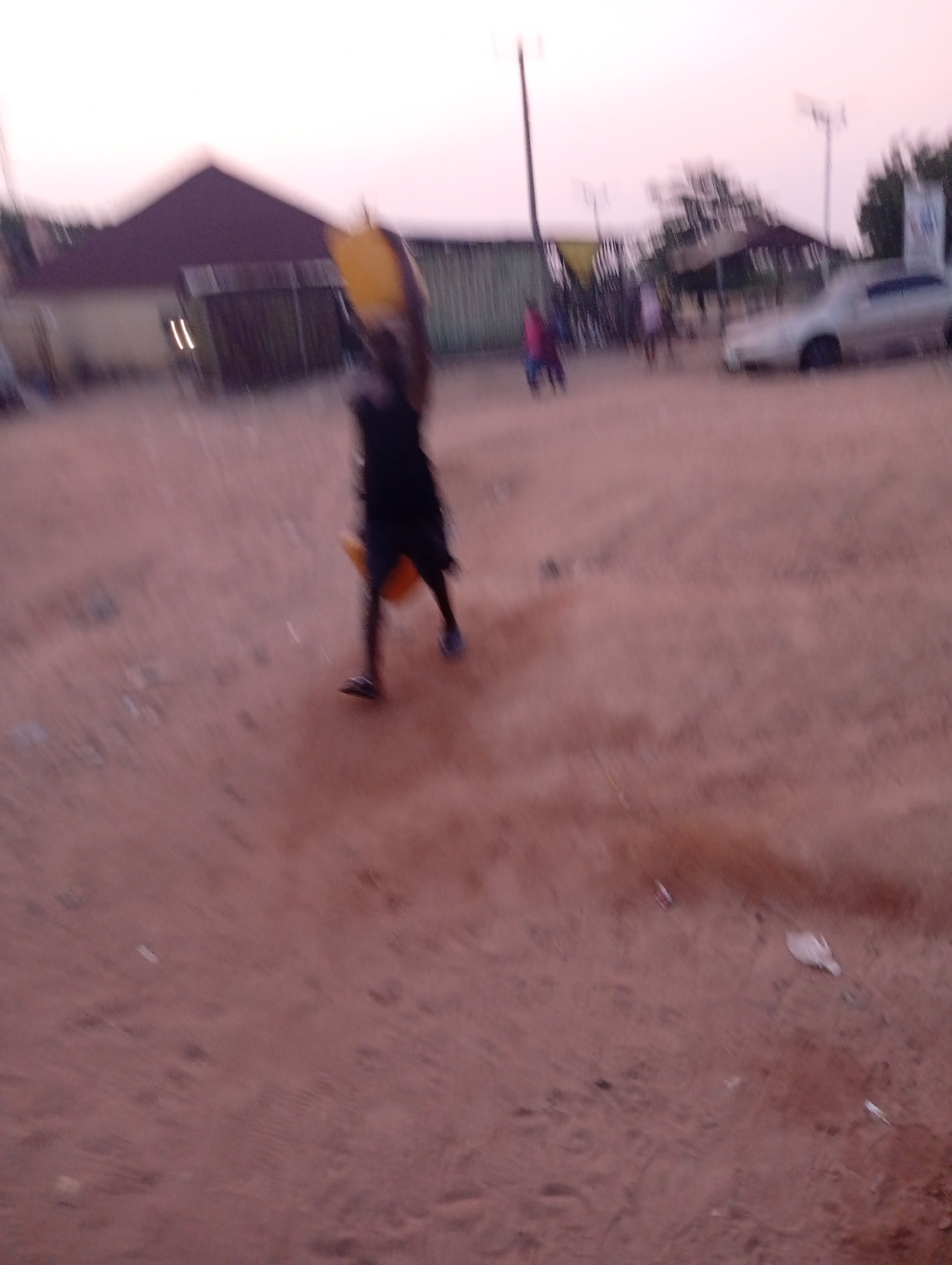 The photograph, taken in Zango-ndaji, Nigeria, depicts a blurred image of an individual carrying a yellow object overhead. The person is wearing dark clothing and sandals. In the background, there are other individuals, a light-colored car, buildings, utility poles, and trees. The ground appears to be sandy or dirt. The sky is a pale, hazy color.