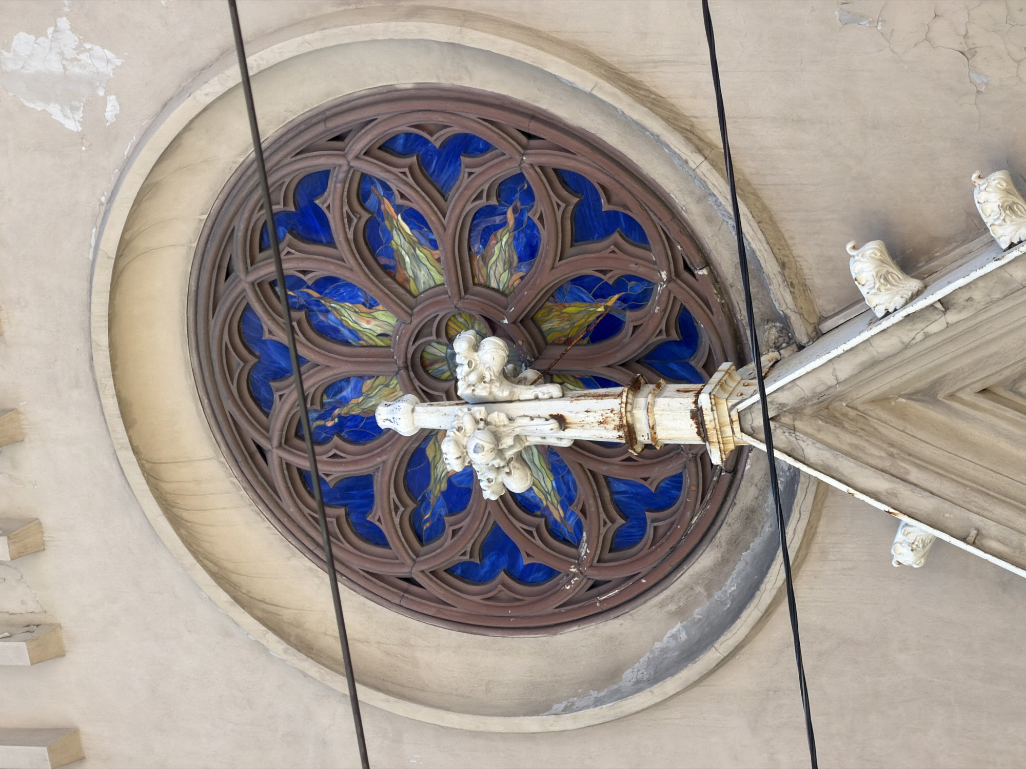 The image shows a close-up of a stained-glass rose window on the exterior of a building, likely a church or historical structure. The window is circular with intricate brown tracery and filled with deep blue glass, interspersed with sections of green and yellow glass that create a floral or flame-like pattern. In the foreground, a weathered white decorative element, possibly part of a cross or spire, extends upwards, showing signs of rust and age. The wall surrounding the window is made of a rough, light-colored stucco with some visible staining and peeling paint, suggesting an older building. Two black electrical wires run horizontally across the frame, partially obscuring the window and the decorative element. The overall impression is one of architectural detail and age. The specific location in Saint Petersburg, Russia, is not evident from the image alone.