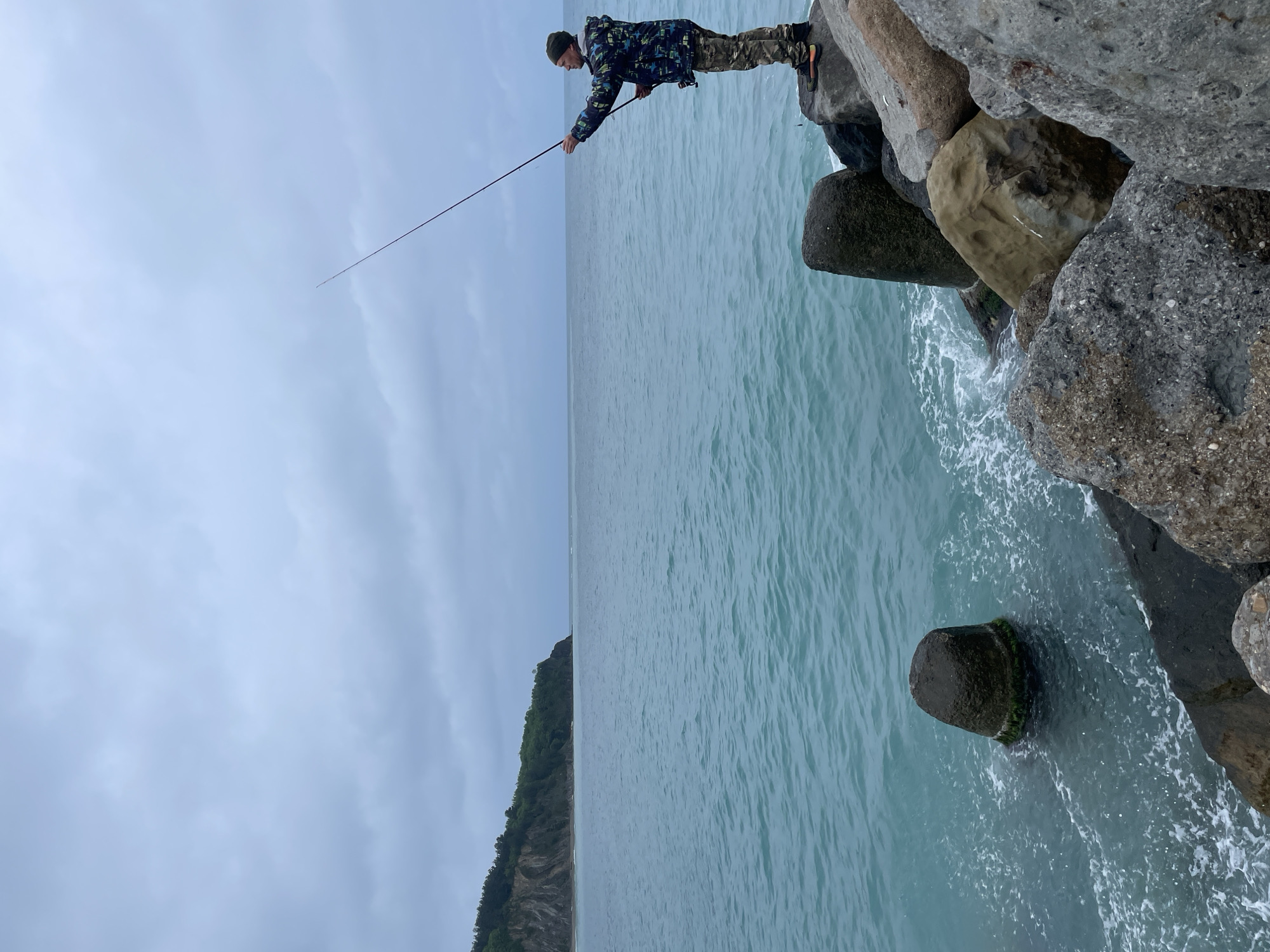 A man stands on large rocks or a breakwater adjacent to the ocean, holding a fishing rod. He is wearing a camouflage jacket, pants, and a dark hat. The ocean is calm with visible waves lapping at the rocks. In the distance, a green, tree-covered landmass is visible under a cloudy sky. The location is possibly the city of Olginka, Russia, implied by the presence of the breakwater and sea.