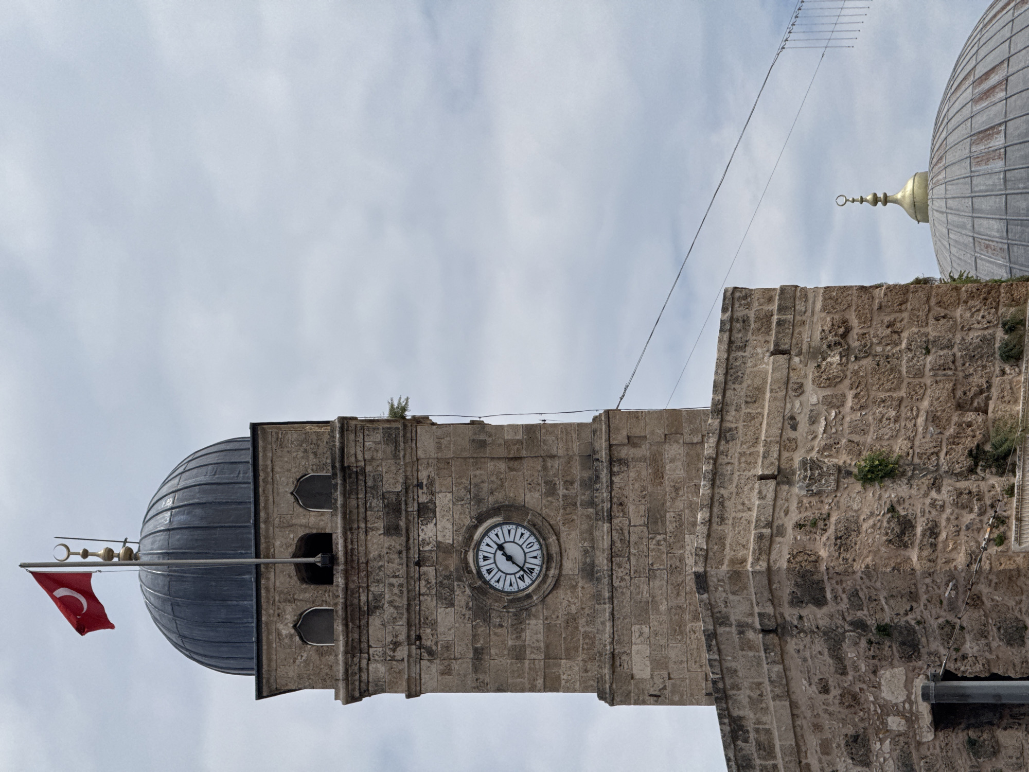 The image depicts a stone tower with a clock face, likely located in Antalya, Turkey. The tower features a dome on top, with a flagpole displaying a Turkish flag. Adjacent to the tower is another building with a dome. An antenna is visible above the buildings against a cloudy sky. The clock on the tower appears to show the time around 8:30. The structures are made of light-colored stone, exhibiting a textured, weathered appearance. No people are visible in the scene.