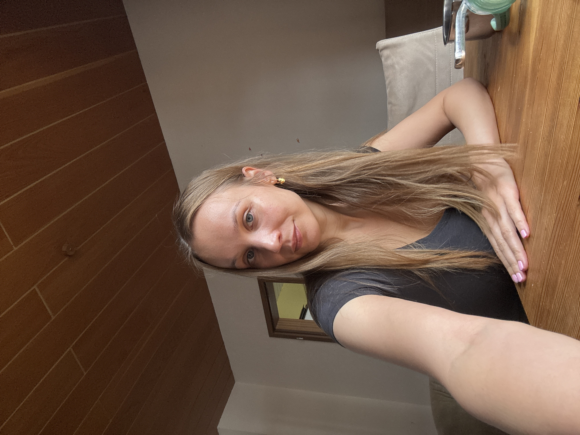 A blonde woman with blue eyes and long, light brown hair is seated at a wooden table, facing the camera. She is wearing a dark gray v-neck t-shirt. She has a small, gold hoop earring in her left ear. Her left arm is resting on the table, with her hand displaying light pink fingernail polish. Behind her, a tan upholstered chair is visible, and a doorway with a yellow-lit interior can be seen in the background. The ceiling is paneled with dark wood. The scene appears to be indoors, within a room in Überherrn, Germany.