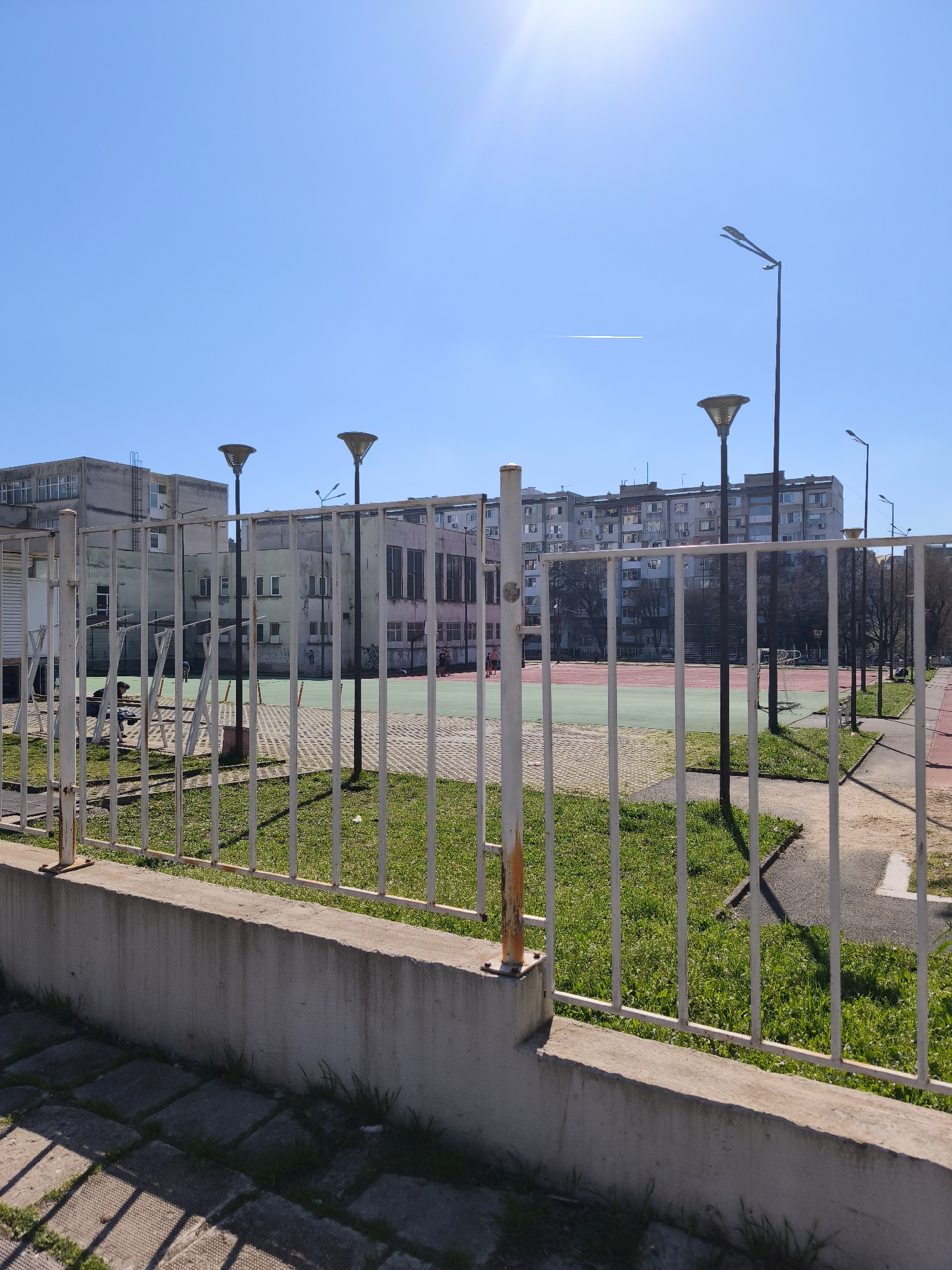 The image depicts a fenced area with a green and red sports court in the background, typical of urban sports facilities. Several buildings are visible in the background with street lights throughout. The area is in Burgas, Bulgaria, and the scene is captured on a clear, sunny day. There are buildings in the distance, showing it is in an urban environment. There is a sidewalk in front of the fence.