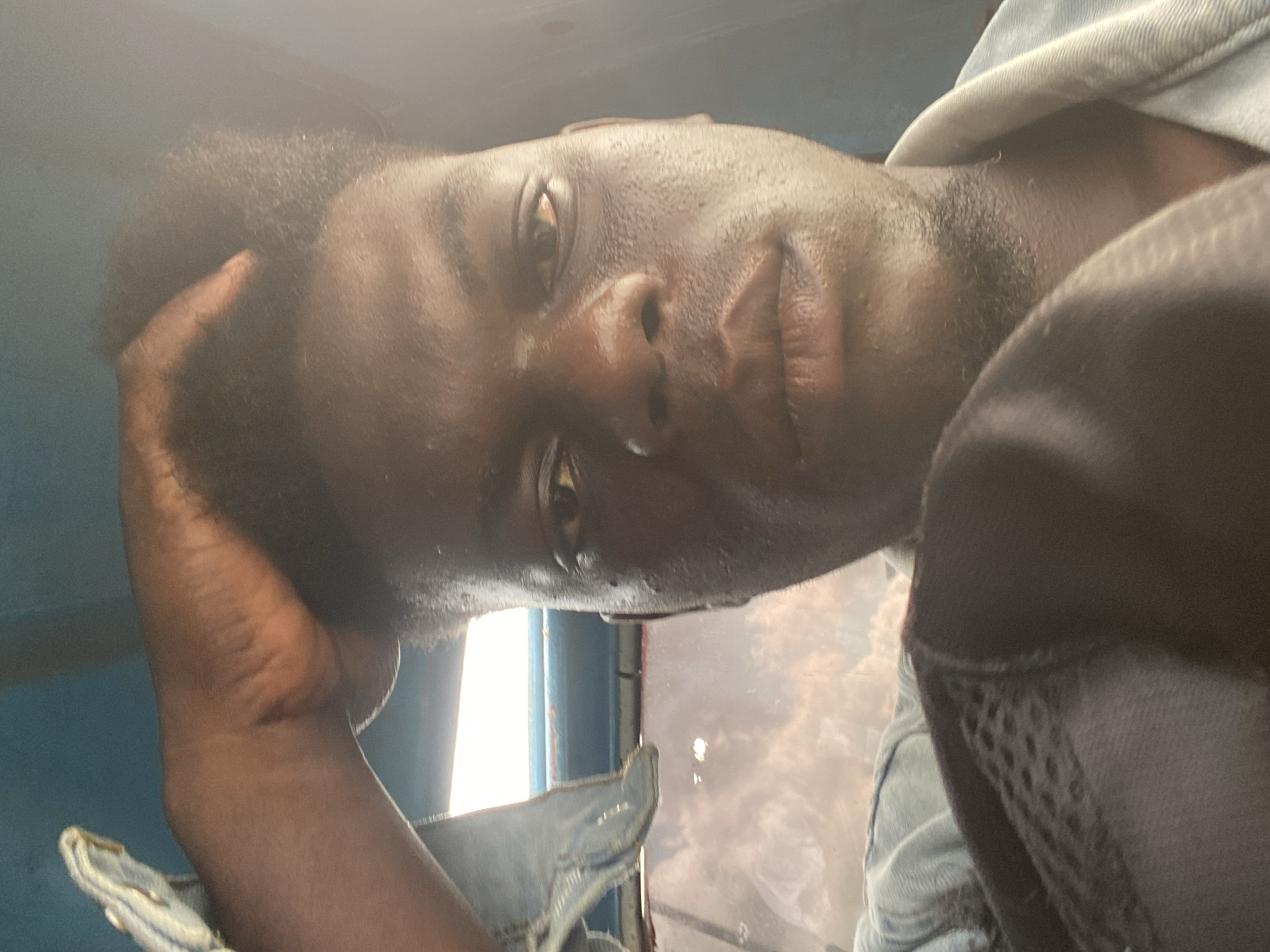 A close-up shot of a dark-skinned person with short, dark hair resting their head on their hand. The person is wearing a light blue denim shirt with a black mesh vest or jacket partially visible on the right side of the frame. The background is partially obscured, but a window with sunlight is visible behind the person. The location is Unknown, Unknown.