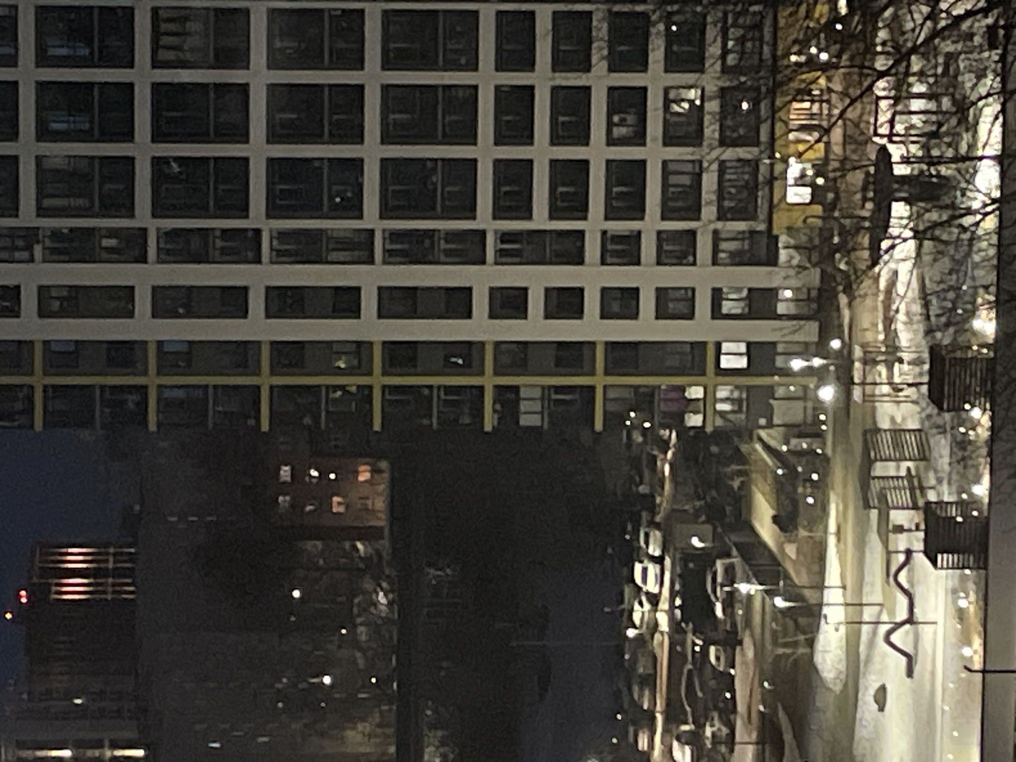 The image shows a view of a multi-story building in Izhevsk, Russia, at night. The building has a grid-like pattern of windows. In the foreground, there is a park with trees and street lights. There are also benches and walkways. Cars are parked in the street alongside the park. Other buildings can be seen in the distance.