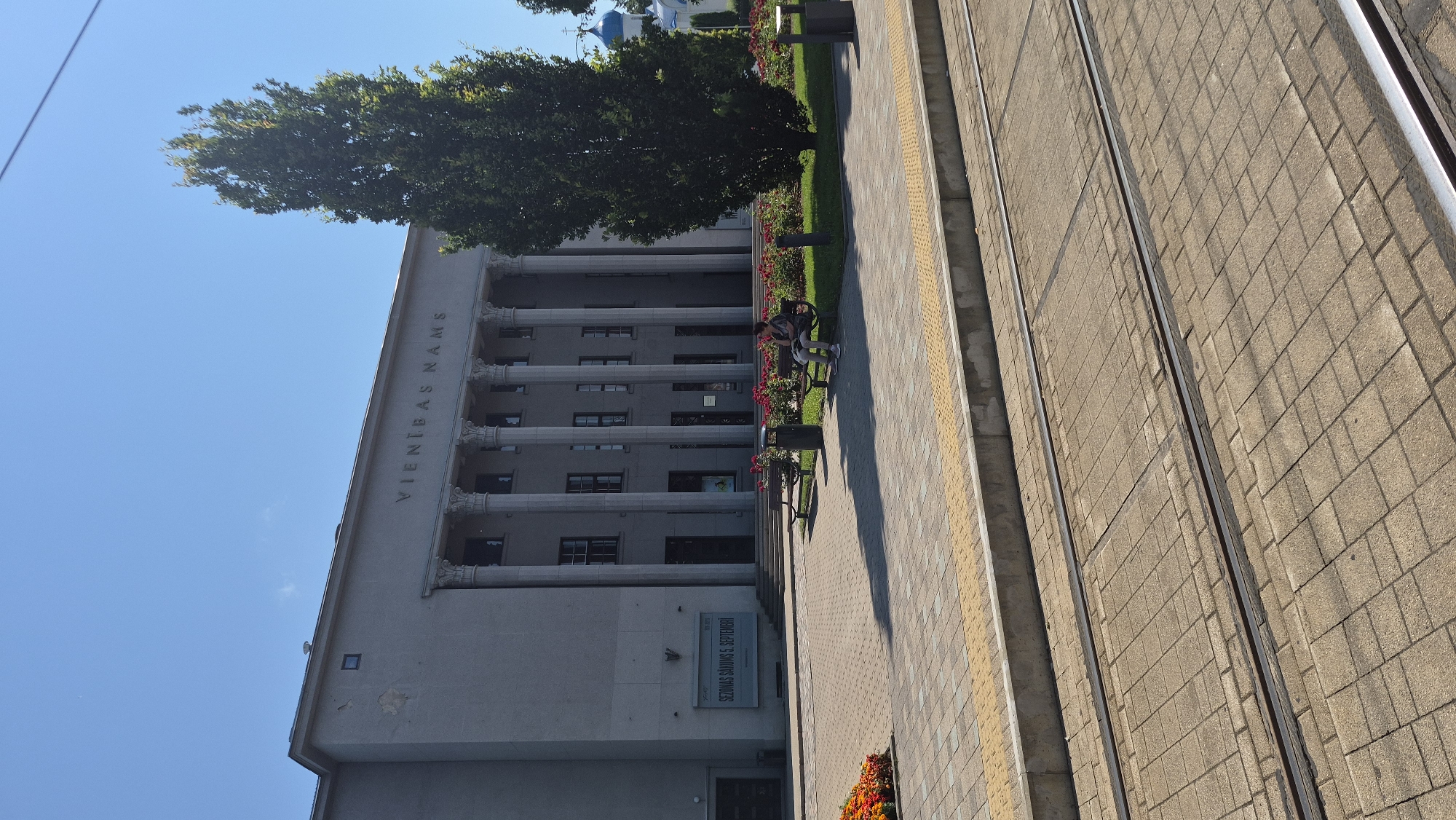 The image shows the "Vienības Nams" (Unity House) in Daugavpils, Latvia, on a clear, sunny day. The building features a neoclassical facade with large columns and a sign above the entrance reading "VIENĪBAS NAMS". In front of the building, there is a paved plaza with tram tracks running through it. A woman is seated on a bench in front of a flower bed, appearing to rest or wait. There are also flower beds with red roses and orange flowers. A tall tree stands to the right of the building. In the background, a building with a blue dome, possibly a church, is partially visible. The overall scene depicts a public space in the city with a mix of architectural styles and natural elements.