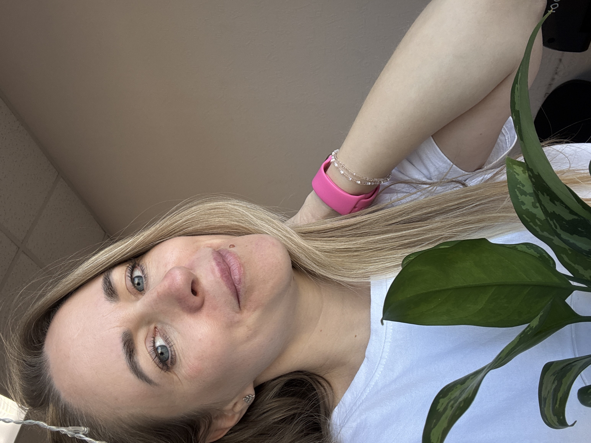 A portrait-style photograph captures a fair-skinned woman with blonde hair and blue eyes. She is wearing a white t-shirt and a bright pink smartwatch with a beaded bracelet on the same wrist. A green leafy plant is partially visible on the right side of the frame. The background appears to be a light beige wall. The location is presumed to be Chaikovsky, Russia.