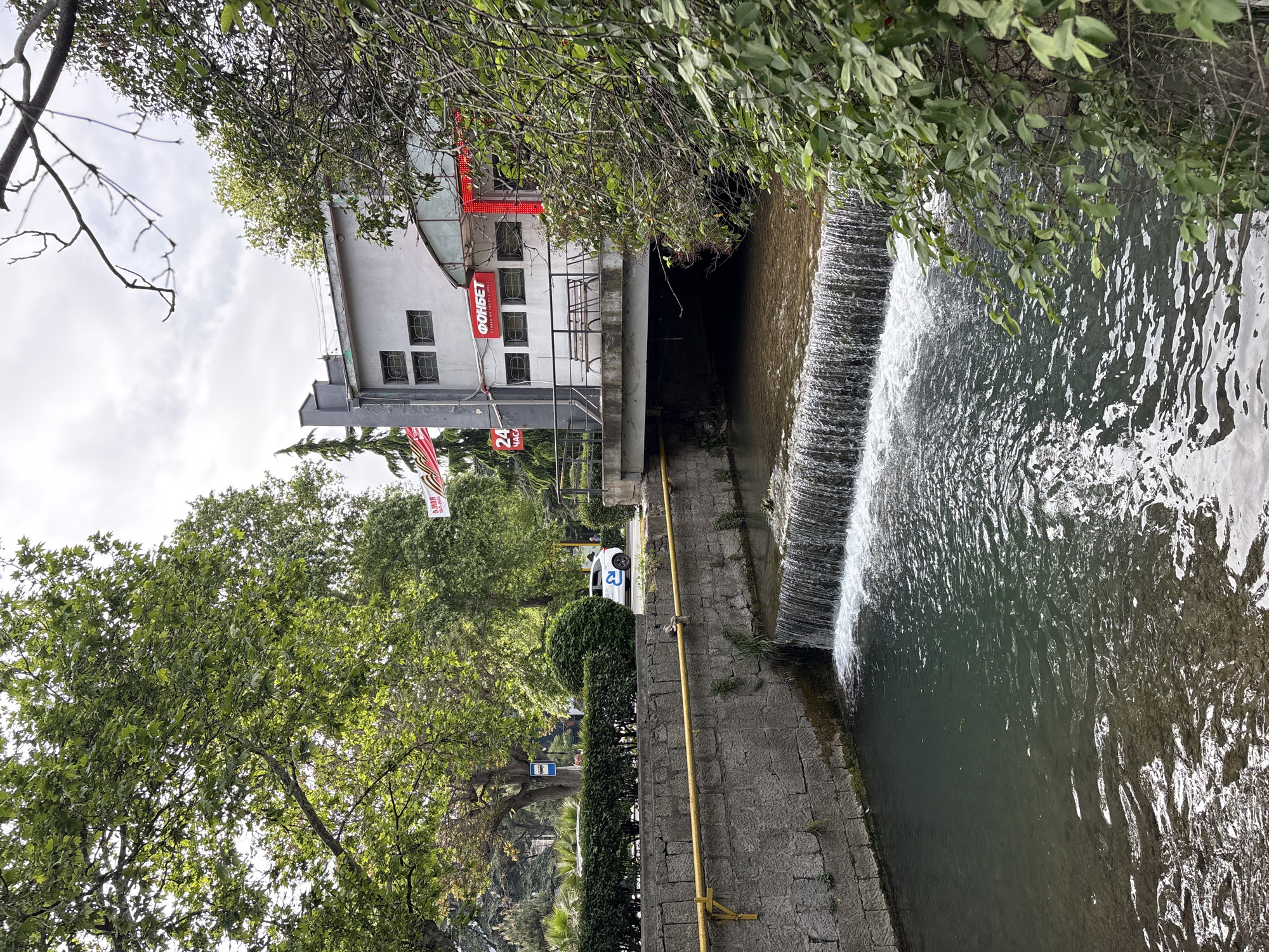 The image depicts a channelized waterway with a small waterfall in город Ялта, Ukraine. A gray building with red signage, including the word "ФОНБЕТ," stands adjacent to the channel. A pedestrian walkway runs alongside the channel, separated by a stone wall and a yellow railing. Trees and greenery frame the scene. A white car is parked on the street. The sky is overcast.