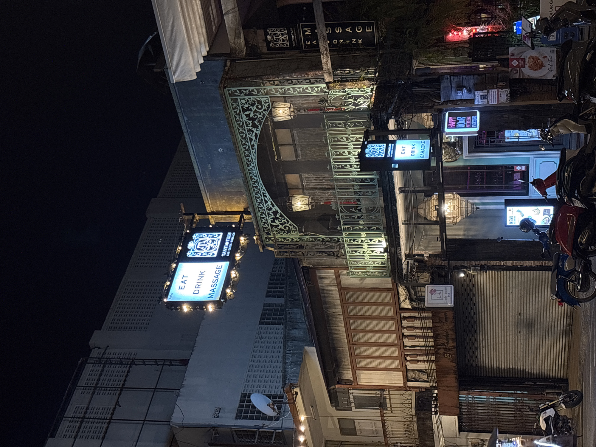 Captured at night in Phuket City Municipality, Thailand, this urban scene features an array of structures and street life elements. An elaborate wrought-iron archway frames a multi-story building displaying signage for "Eat, Drink, Massage" and "Eat, Drink, Karaoke." Adjacent buildings show rows of windows and closed shutter doors. A parked motorcycle and scooters are seen on the street. Various illuminated signs indicate commercial establishments, including one for “Happy Hour”. The lighting is a mix of electric signage, streetlights, and illumination from the businesses.