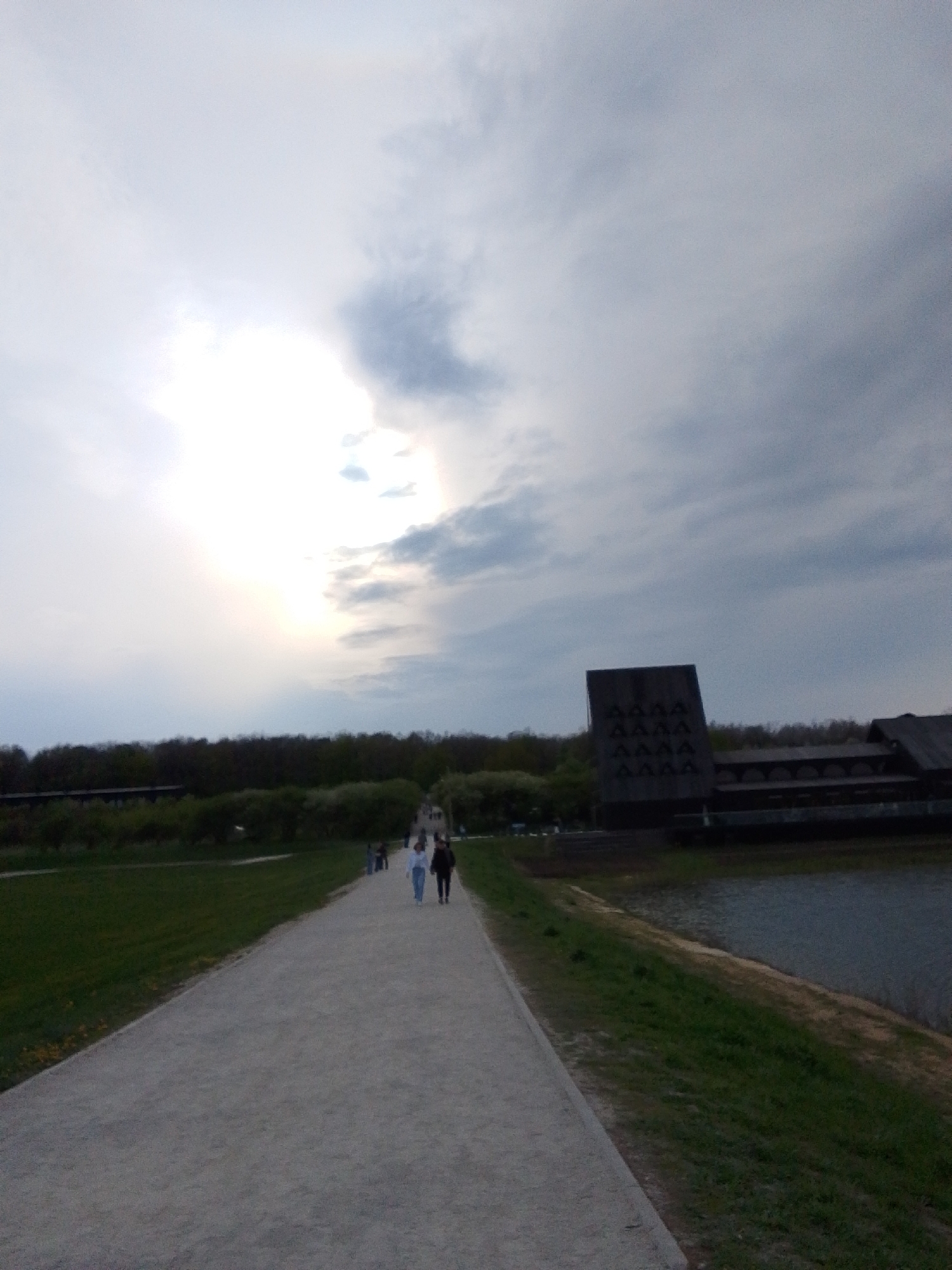 The image shows a gravel path in a park in Алисово, Russia. Two people are walking on the path away from the camera. To the right of the path is a body of water. Beyond the water is a dark building with a decorative facade. To the left of the path is a grassy area, followed by a line of trees. The sky is overcast with the sun shining through the clouds.