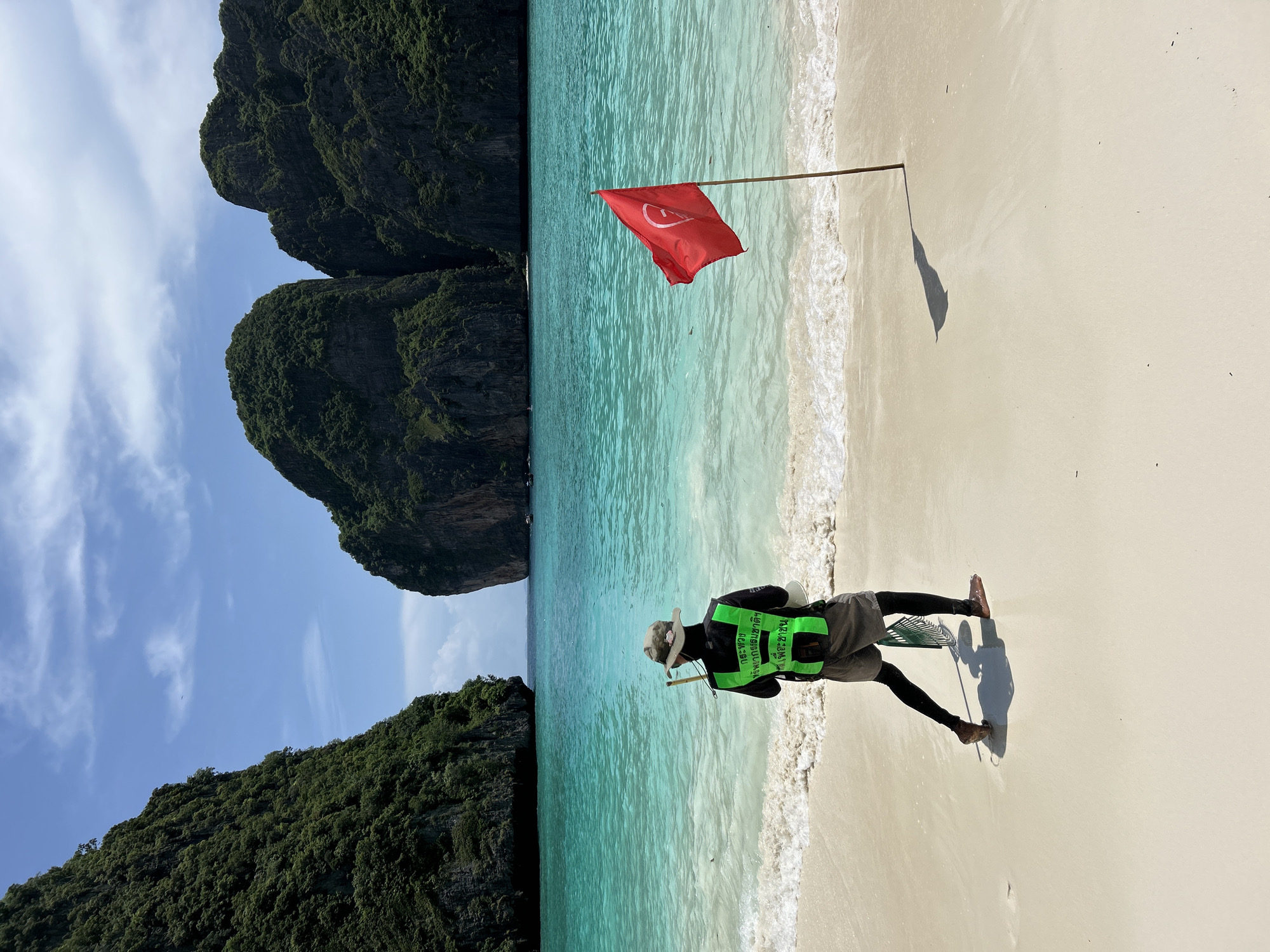 The image depicts a scenic view of Maya Bay, located in the Phi Phi Islands, near Ao Nang, Thailand. The bay is characterized by its turquoise waters, white sandy beach, and towering limestone cliffs covered in lush greenery. In the foreground, a person wearing a hat, a green reflective vest with Thai text, and dark leggings stands facing the water. They are holding a stick and a net-like bag. A red flag with a white symbol is planted in the sand to the right of the person, its shadow stretching across the beach. The water exhibits gentle waves lapping at the shore, and the sky is a clear blue with scattered white clouds. Several boats can be seen in the distance, suggesting the bay is a popular tourist destination.