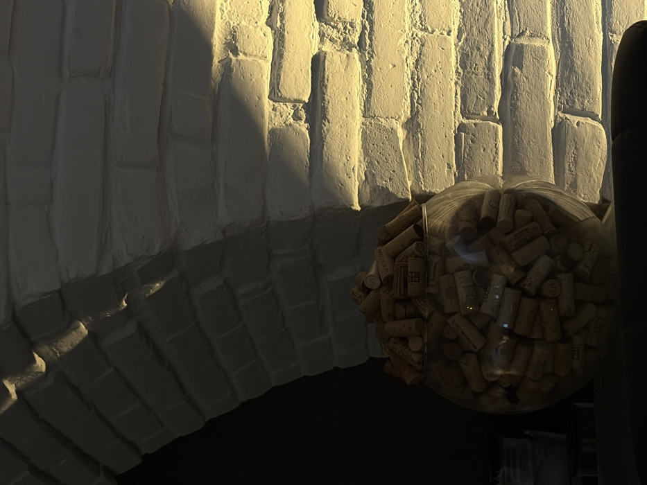 A curved, white brick wall is illuminated by a warm light source. The light casts shadows that accentuate the individual bricks. In the foreground, a clear glass container is filled with numerous wine corks. A dark vertical object is visible to the right of the container. The lower portion of the scene is consumed by deep shadows, creating a stark contrast.