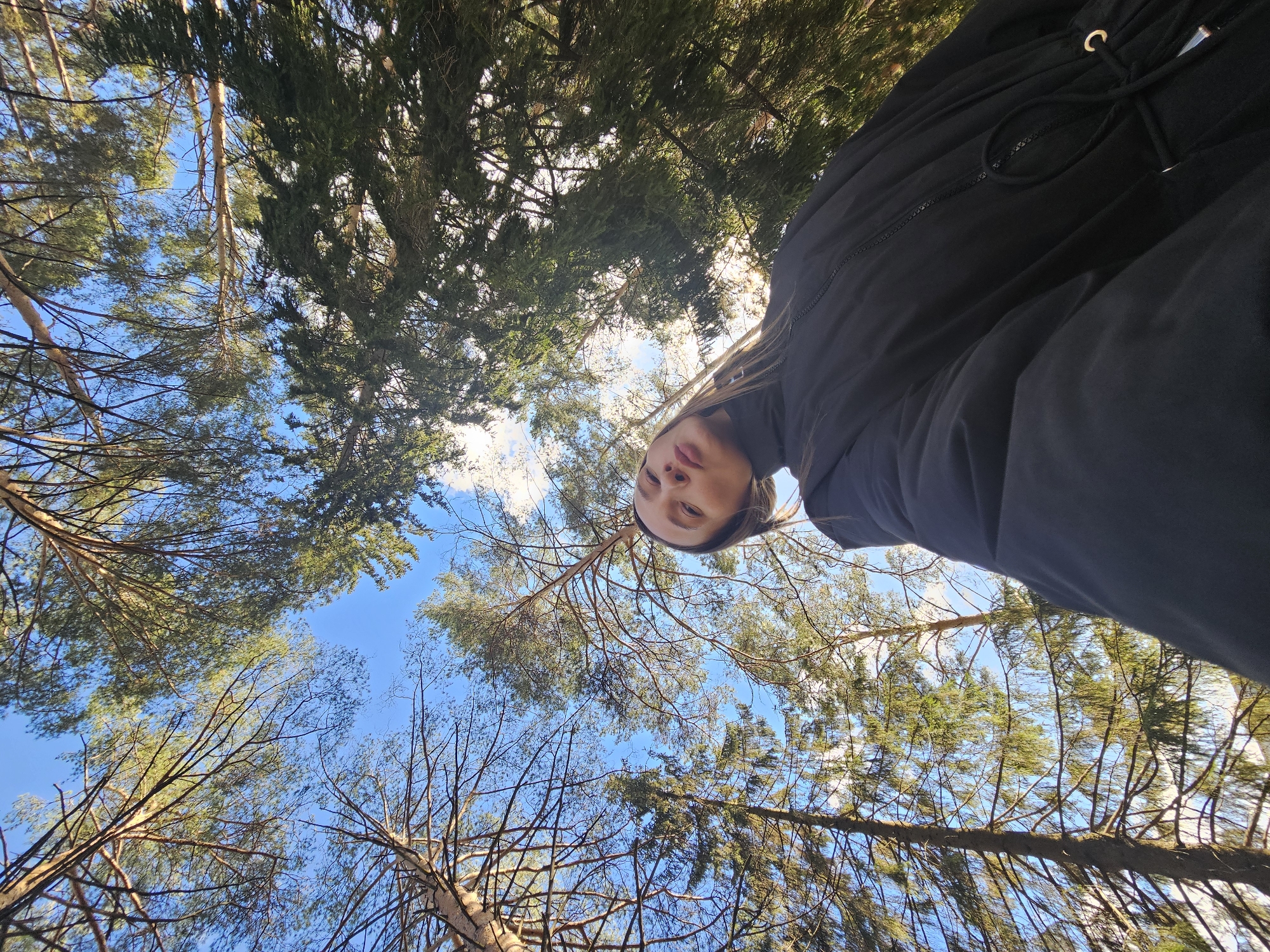 A low-angle photograph captures a woman looking down at the camera, framed by a canopy of trees against a blue sky. The woman wears a black jacket with drawstring details. The trees are a mix of coniferous and deciduous types. Some trees are bare-branched, while others are full of green foliage. The bright sky is visible through the gaps in the tree branches. The photo was taken outdoors in a natural setting, with the perspective suggesting the photographer was lying on the ground. The city of Chaikovsky, Russia, as the location, is implied.