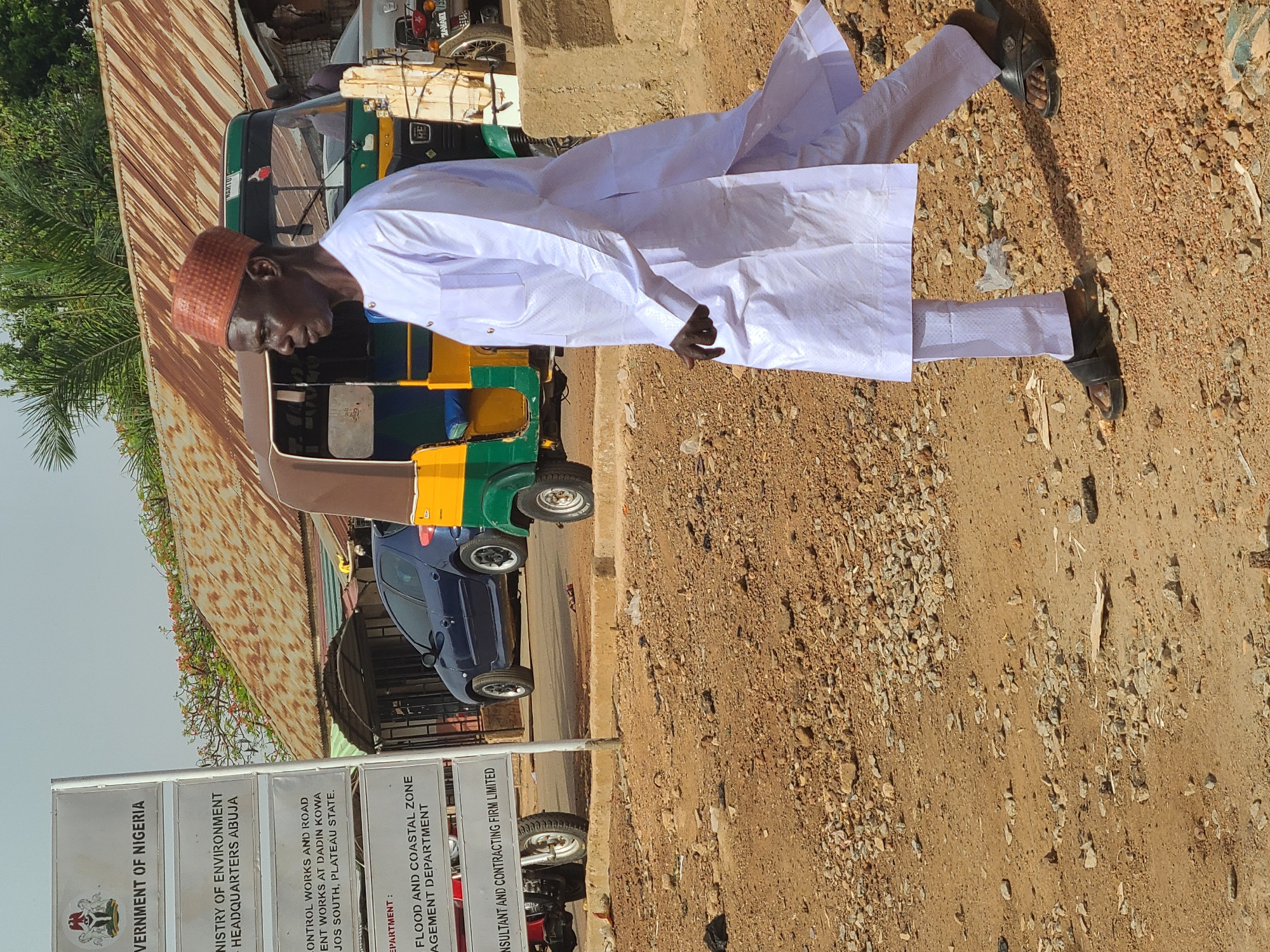 A full-length image shows an African man wearing traditional white attire with a brown woven cap, standing on a dirt road beside a green and yellow auto-rickshaw. The man is facing left, his arms at his side. He's wearing sandals. Behind him is a sign that reads "Government of Nigeria, Ministry of Environment Headquarters Abuja" and includes control works and road maintenance information for Jos South, Plateau State. A blue vehicle is also parked nearby. The background includes a corrugated metal roof and sparse vegetation. The setting suggests a rural or semi-urban area within Jos, Nigeria.