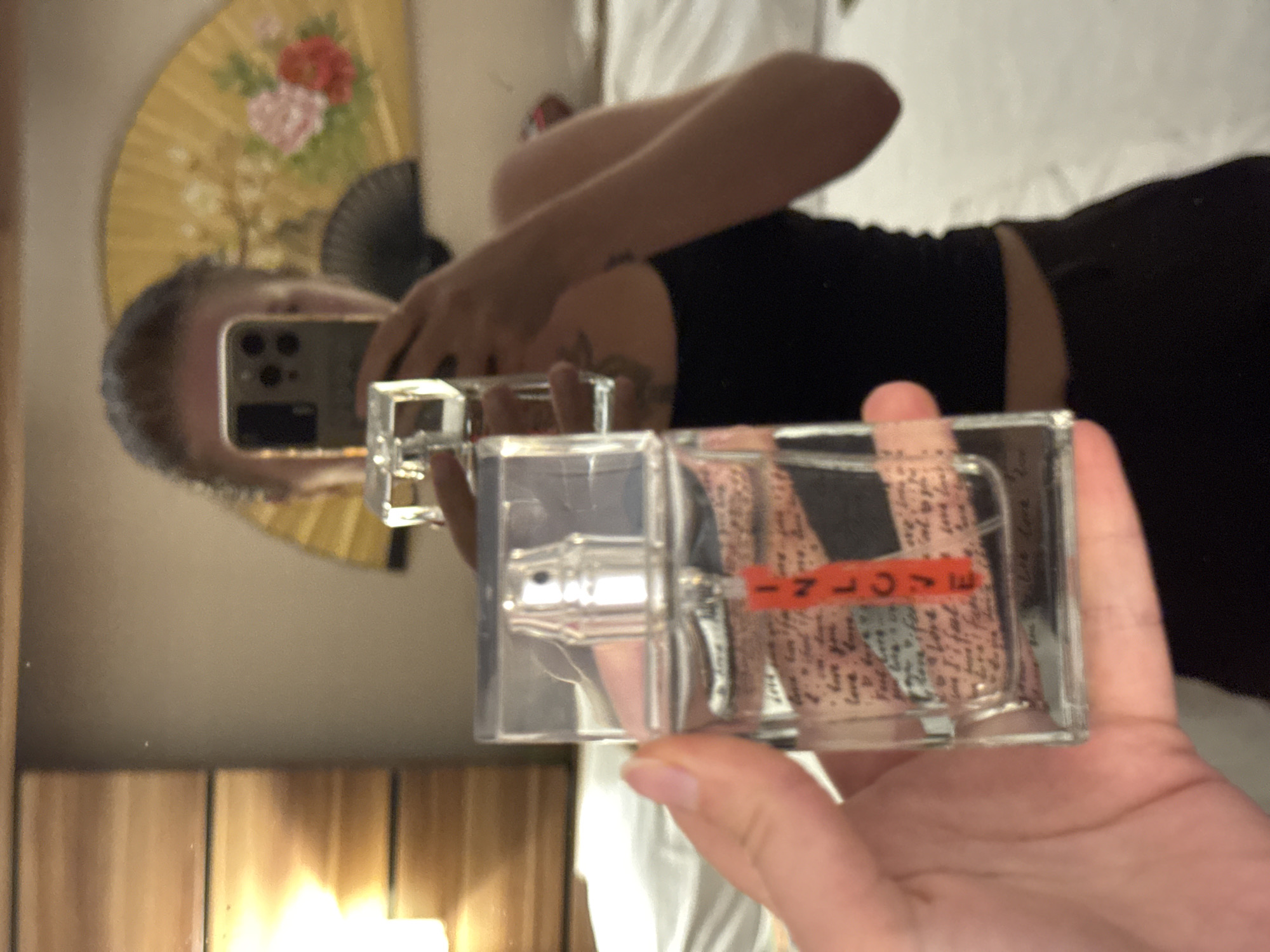 A person's hand is holding a clear glass perfume bottle in the foreground. The bottle has a silver spray nozzle and a clear cap. Inside the bottle, there is a clear liquid and a label with text and an orange stripe that spells out "INLOVE". The background is a blurry reflection in a mirror, showing a woman holding a smartphone, a yellow fan with pink and red flowers, and a wooden headboard with a light. The setting appears to be indoors, possibly a hotel room or bedroom.