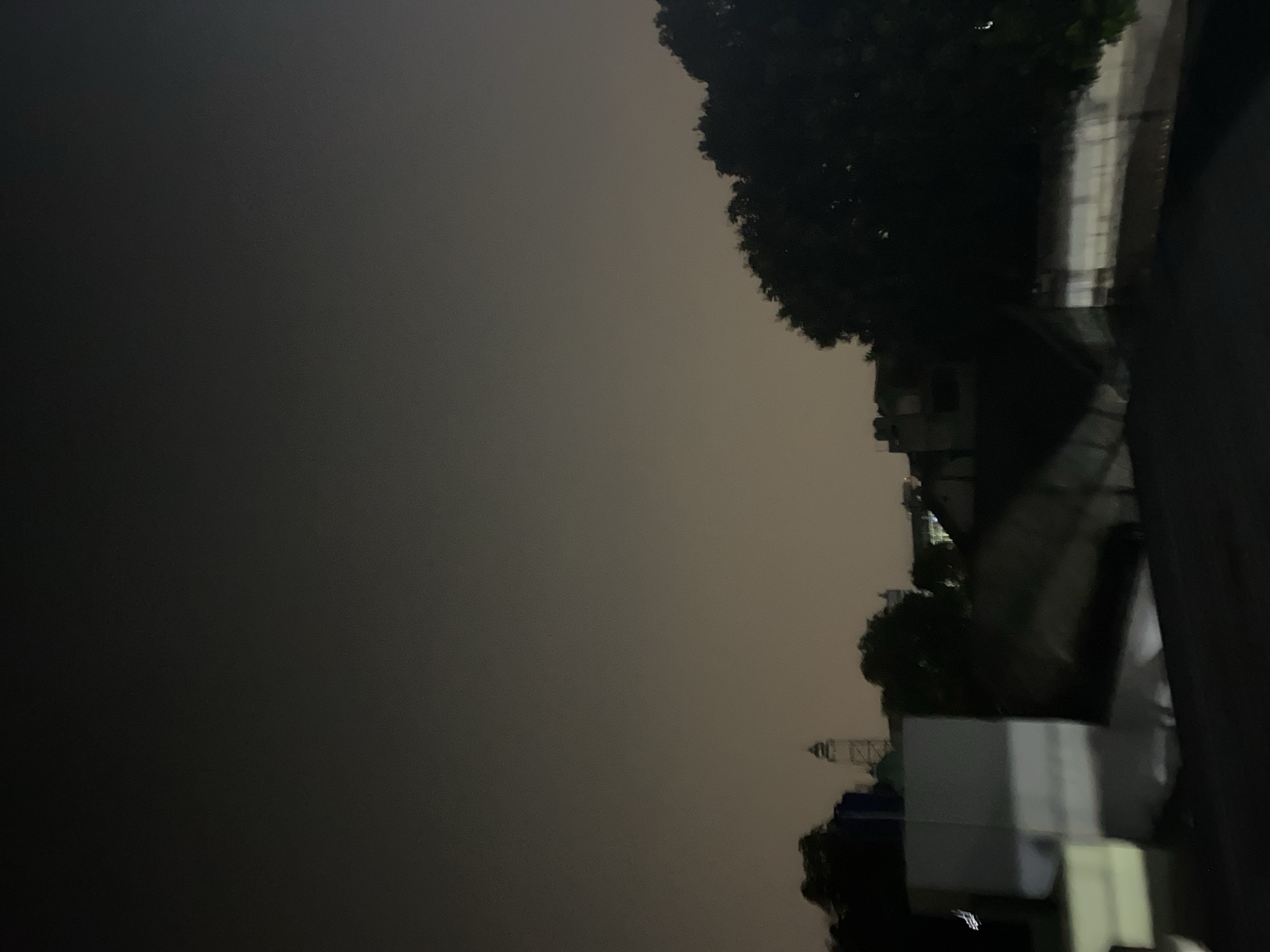 This is a nighttime photograph of a cityscape. In the foreground, there are dark, indistinct structures, possibly rooftops, with some illuminated elements. In the mid-ground, there are more buildings and trees. A notable structure is a tower or spire, which appears to be a prominent landmark. The sky is overcast and dark. The overall impression is of a quiet urban scene at night, possibly in a residential or mixed-use area. Based on the provided hints, the scene is located in the city of Tangerang, Indonesia.