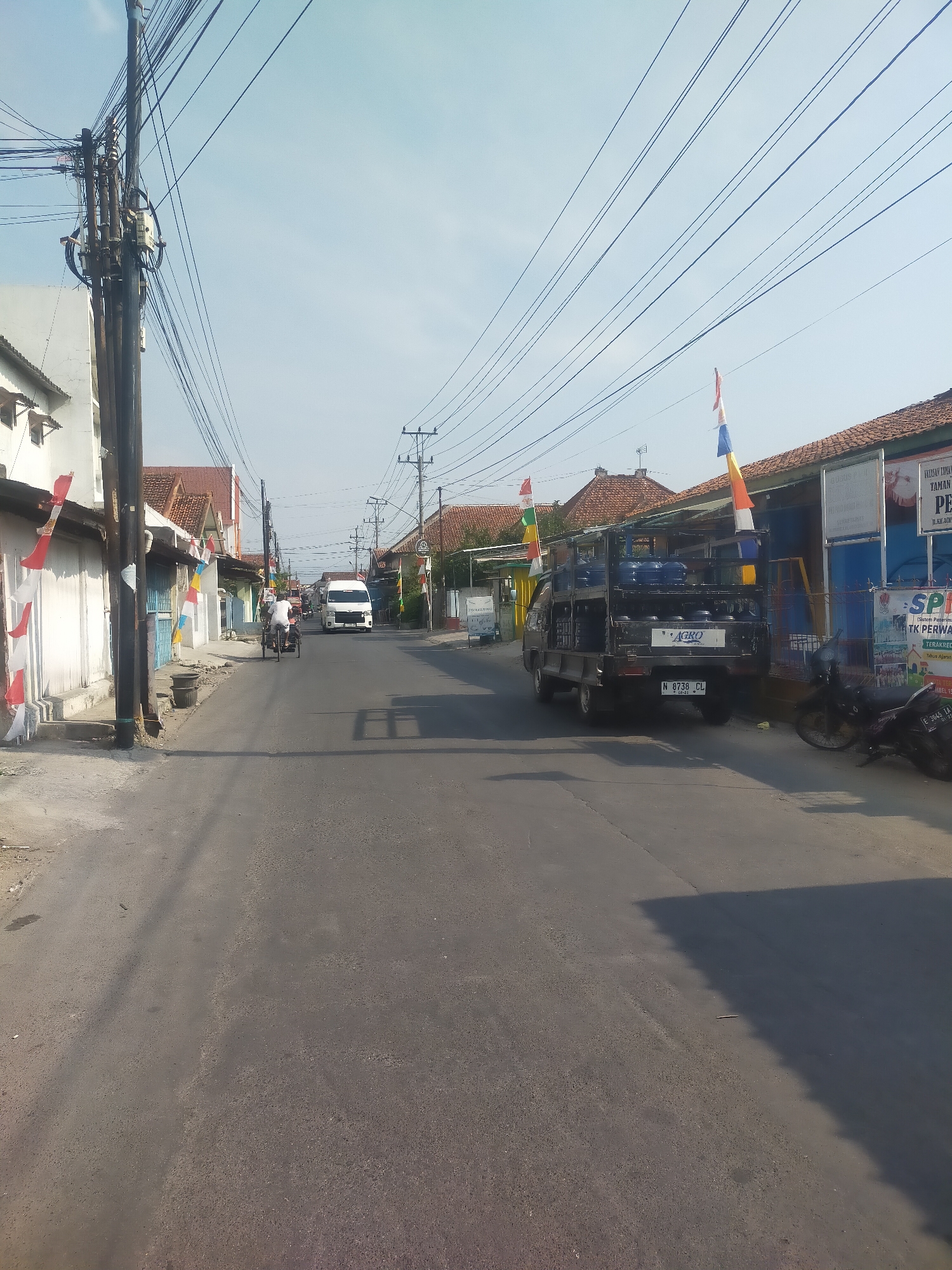 The image depicts a street scene in Slawi Kulon, Indonesia, under a clear blue sky. The street is paved and appears to be a two-lane road with parked vehicles and buildings lining both sides. Prominently featured on the right side of the street is a dark-colored pickup truck loaded with blue water containers. The truck has a license plate displaying "N 8738 CL". Further down the street, a white van is visible.
On the left side of the street, a white building with red-tiled roofs is partially visible, adorned with red and white Indonesian flags. A man on a bicycle with a passenger is seen riding away from the viewer on the left side of the road, and another bicycle is parked nearby. Power lines are strung across the scene, supported by utility poles.
The environment suggests a typical urban or suburban setting in Indonesia. The overall impression is one of a sunny day with moderate activity on the street.