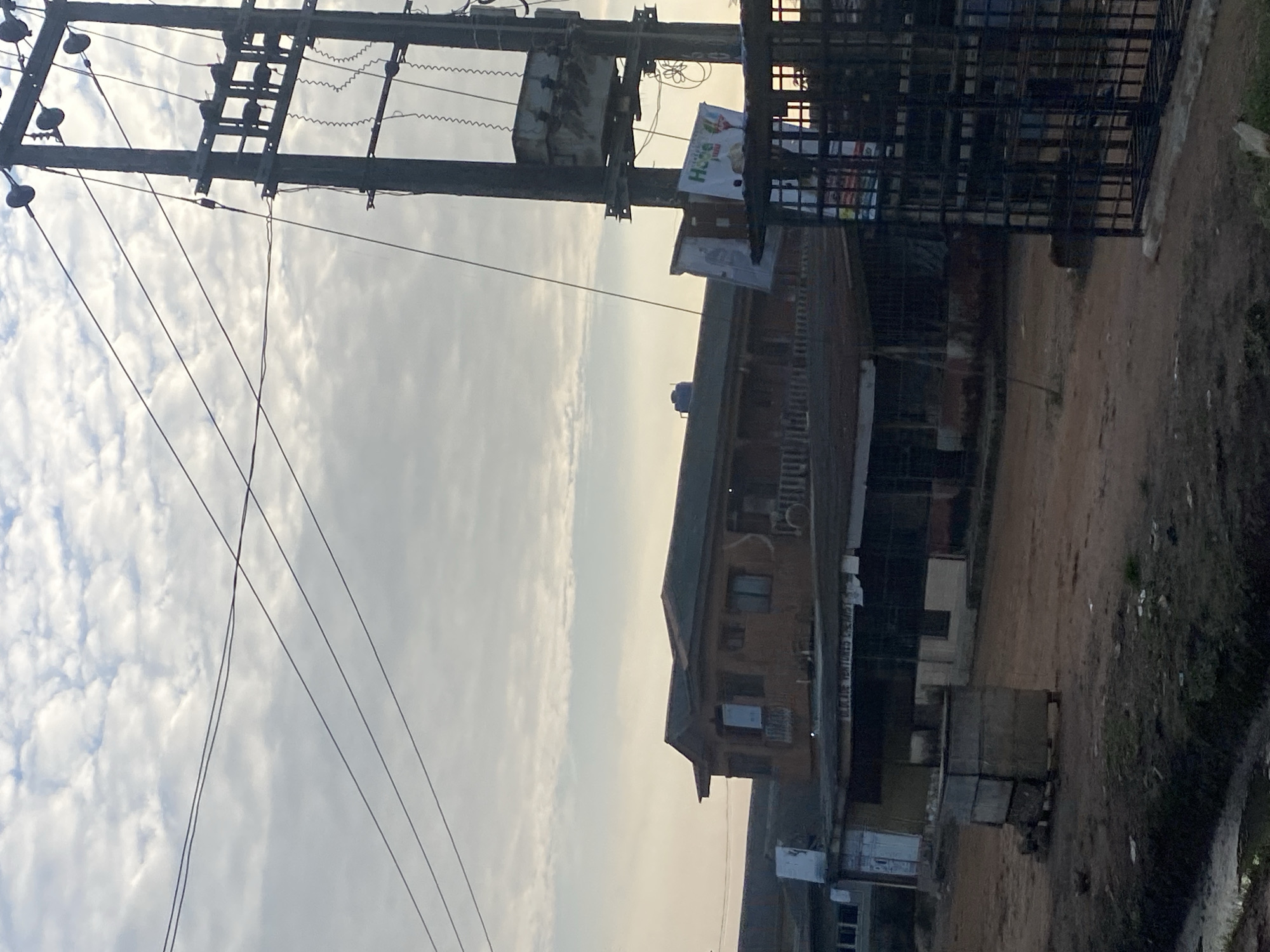 In Ikorodu, Nigeria, a street scene unfolds under a cloudy sky. Dominating the right side of the frame is a tall, dark utility pole with a transformer attached. Multiple electrical wires stretch across the image, some taut and others slack, creating a network against the sky. In the midground, a two-story building with a brown facade and a darker roof stands prominently. The lower level appears to be a series of shops or businesses, with awnings and openings visible. A sign reads "OLAKINE VENTURES CHEMIST" above one of these establishments. To the left of this building, another structure with a white front is partially visible. In the foreground, the ground is a mix of dirt and mud, with some scattered debris. A large, grey concrete structure, possibly a bin or a planter, sits on the left side of the dirt road. A black metal gate or fence encloses an area on the right side of the image, obscuring some of the businesses behind it. A few small items, such as what appear to be stones or bricks, are placed near the concrete structure. The overall impression is one of a functional, lived-in urban environment.
