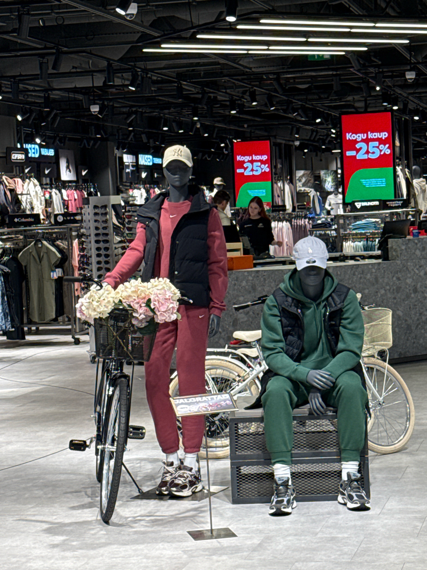 The image depicts the interior of a retail store, likely a sportswear or fashion outlet, in Tallinn, Estonia. Two mannequins are prominently displayed. The left mannequin, dressed in a burgundy tracksuit and a black puffer vest, stands next to a bicycle with a basket of flowers. The right mannequin, dressed in a green tracksuit and a black vest, is seated on a metal crate. In the background, clothing racks are visible, and large digital screens display "Kogu kaup -25%" (meaning "Entire stock -25%") in red and green colors, indicating a sale. The store features industrial-style lighting and shelving.