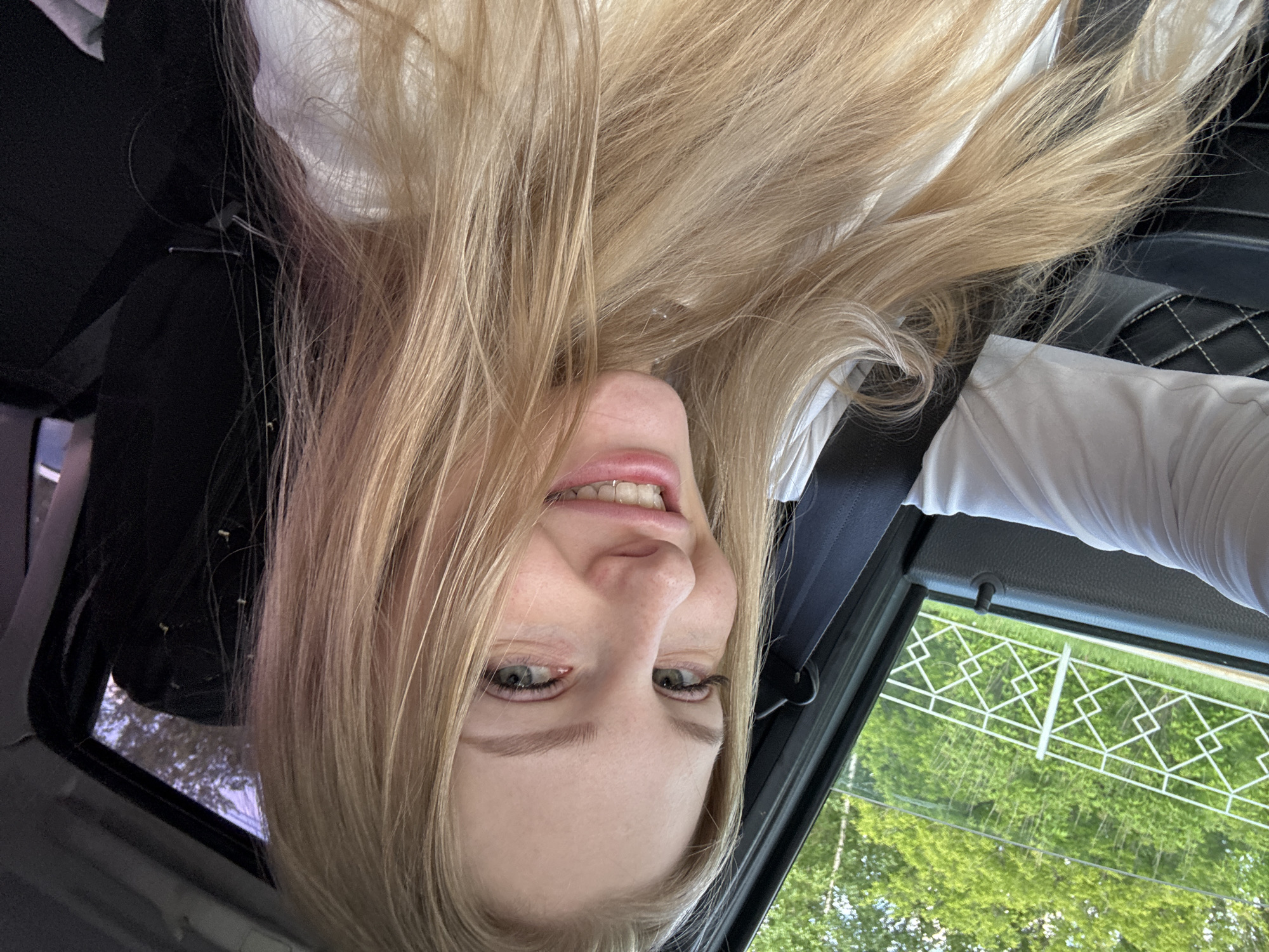 An overhead shot captured inside a vehicle in Perm, Russia, features a young woman with long, straight, blonde hair, smiling playfully directly into the camera. Her face is presented upside down in the frame, with her blonde hair cascading down around her head. She has light-colored eyes and a wide smile, revealing braces on her upper teeth. She appears to be wearing a white shirt or blouse.
The setting is clearly inside a car, with elements of the interior visible: a dark car seat with quilted stitching on the right, and what appears to be a black headrest or part of the car's interior on the left. Through the car window, a bright outdoor scene is visible, suggesting daytime. Lush green foliage, likely trees, fills the background. A distinctive white fence or railing with a repeating geometric, diamond-shaped pattern is reflected or positioned outside the window, adding a specific architectural detail to the outdoor environment. The bright lighting and vibrant green indicate a clear, possibly sunny, day. No text is visible in the image.