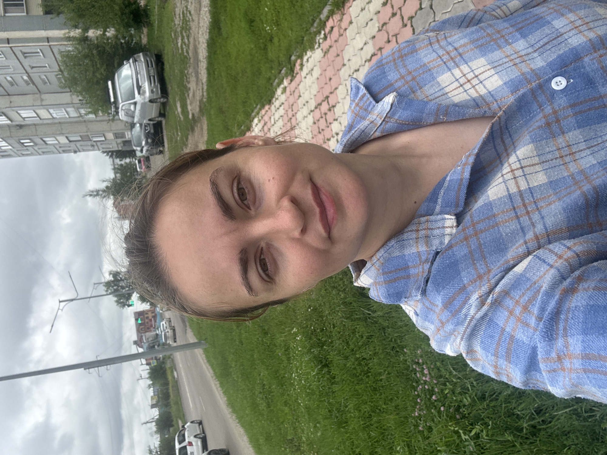 A woman is taking a selfie outdoors in Blagoveshchensk, Russia. She is wearing a blue and white plaid shirt. Behind her, there is a sidewalk with a pink and white patterned walkway, green grass, a street with cars, and a tall, grey apartment building. The sky is cloudy.