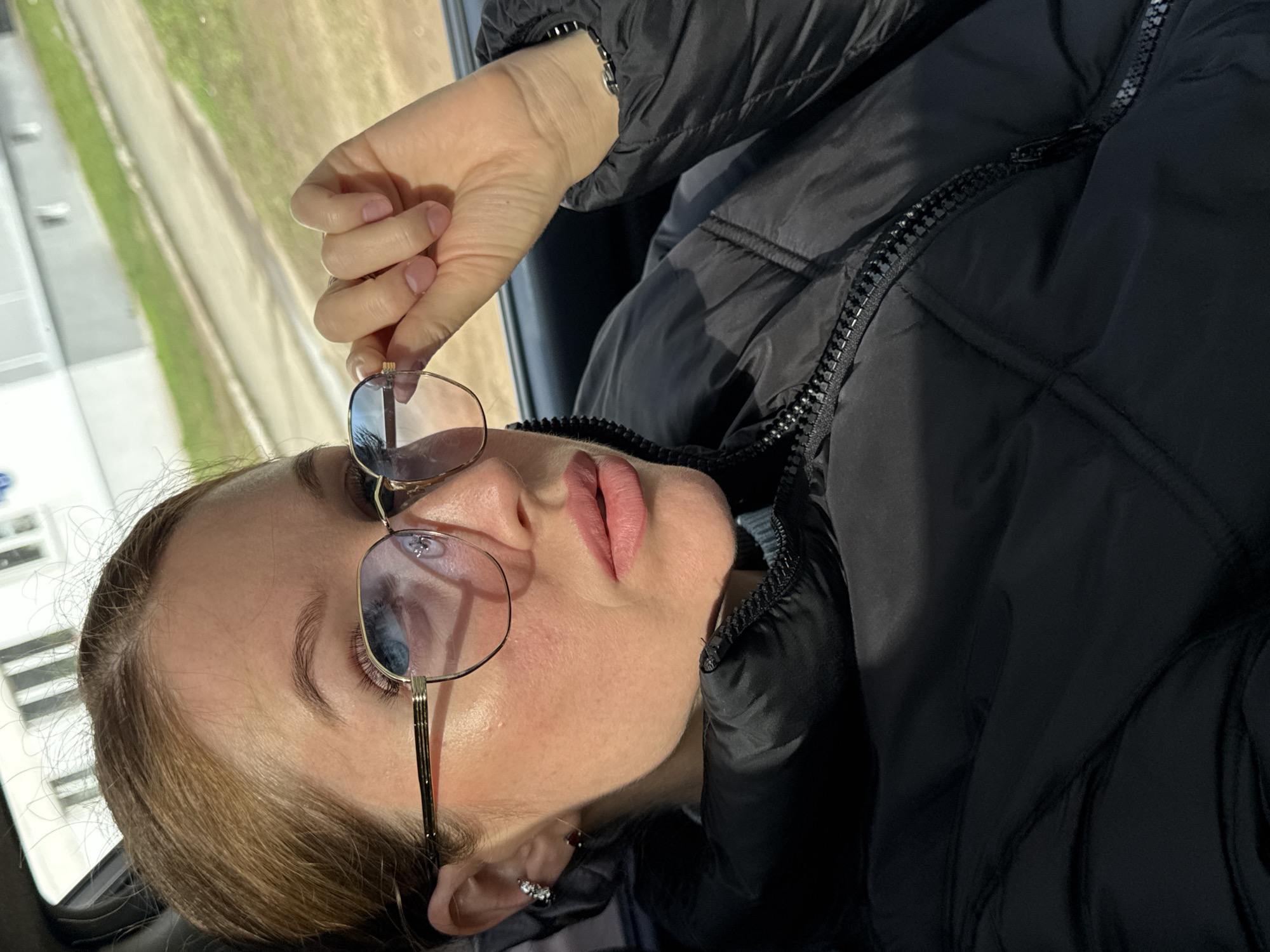 The image is a portrait of a young woman, taken from a slightly low angle, while she is inside a vehicle in Chaikovsky, Russia. She has light brown hair, blue eyes, and pinkish lips, with a neutral to slightly contemplative expression. She wears gold-framed eyeglasses, which she is holding by the bridge with her right hand, revealing a ring on her finger. A silver earring with multiple small stones is visible on her left earlobe, accompanied by another darker stud. She is dressed in a black quilted or puffer jacket, which is zipped up, indicating cool weather.
The setting appears to be inside a car or bus, with an outdoor scene visible and blurred through a window in the background. This background suggests bright daylight, possibly sunny weather, showing a light-colored path or road with green strips of grass or fields on either side. Reflections in her eyeglasses further indicate a bright outdoor environment, displaying patches of blue sky and hints of distant structures. No visible text is present in the image.