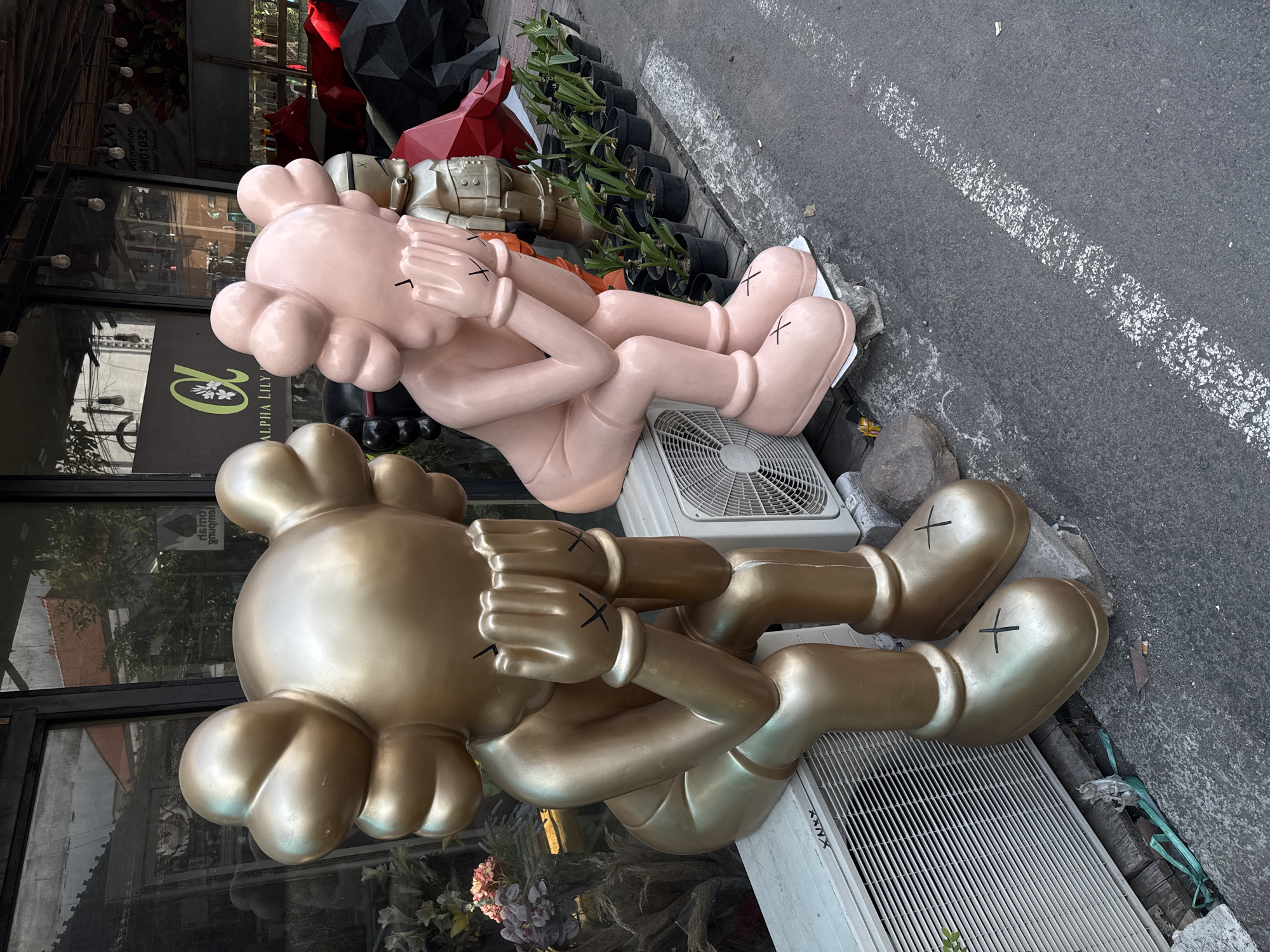 The image shows two large, cartoonish figures, one gold and one pink, sitting on top of air conditioning units on a sidewalk in North Kuta, Indonesia. Both figures are in a pensive pose, with their heads bowed and hands covering their faces, depicting a common pose of distress or contemplation. The figures are made of a smooth, matte material and have distinctive "X" marks on their gloved hands and large, round feet.
In the background, there is a storefront with large windows, reflecting the surroundings. A sign is visible above the window, displaying the text "ALPHA LILY" and a green logo. Various plants in black pots are arranged on wooden planks behind the figures, suggesting a commercial or retail setting, possibly a flower shop or nursery. The sidewalk is made of asphalt, with a white line painted on it, indicating a parking or traffic boundary. The overall scene is outdoors, under natural lighting.