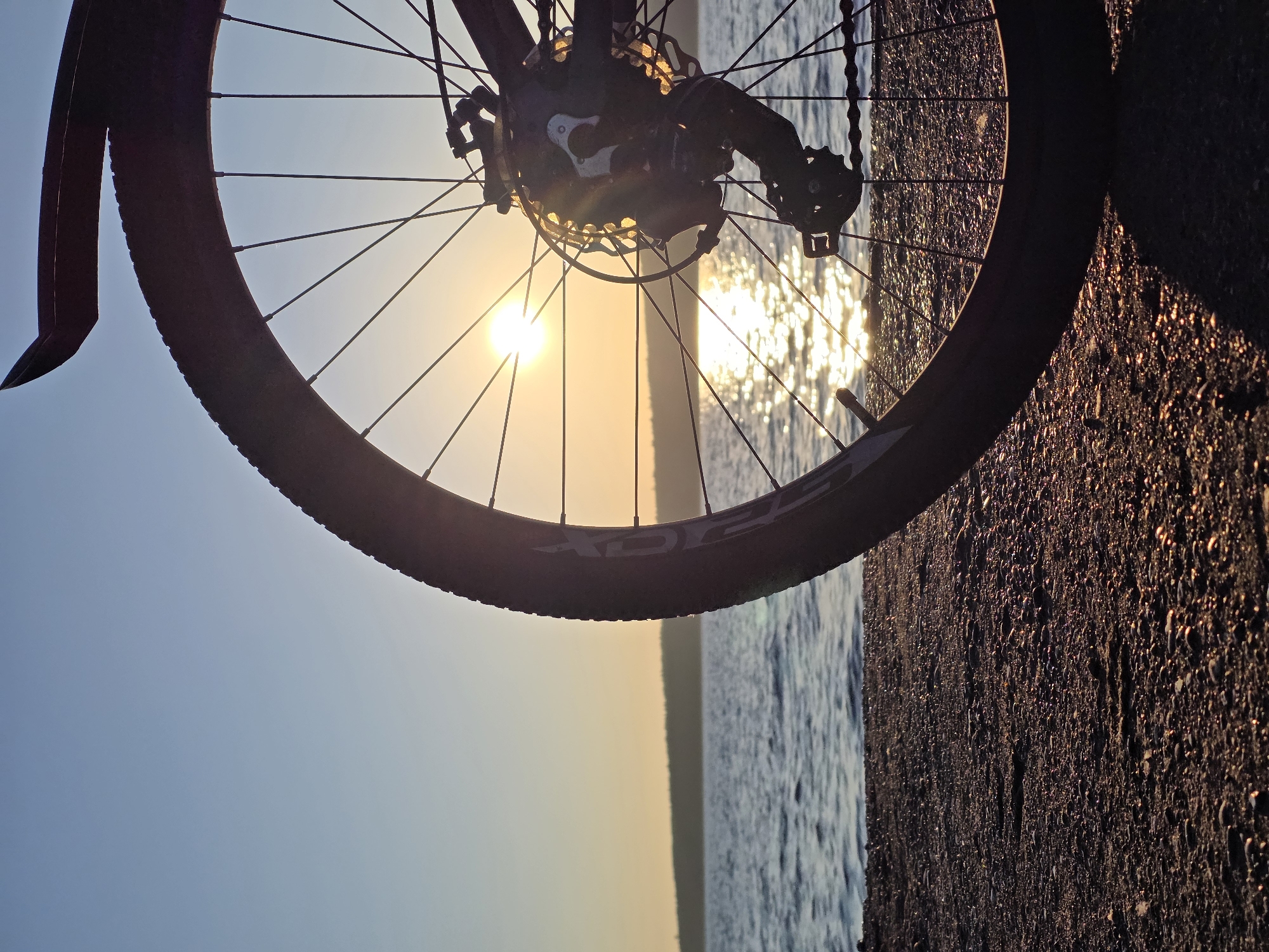 The image shows a close-up of a bicycle wheel parked on a rocky shore, with the sun setting over a body of water. The sun is visible in the background, casting a golden reflection on the water. The bicycle wheel is in the foreground, obscuring part of the view. The terrain is covered with small pebbles and stones. The water appears calm with gentle ripples. In the distance, a treeline can be seen along the horizon. The sky is a clear gradient of blue and yellow. The scene is suggestive of a serene outdoor setting, possibly near a lake or river in the city of Chaikovsky, Russia, given the characteristic landscape. There are no people or animals present in the image. The focus is on the interplay of light, water, and the silhouette of the bicycle against the sunset.
