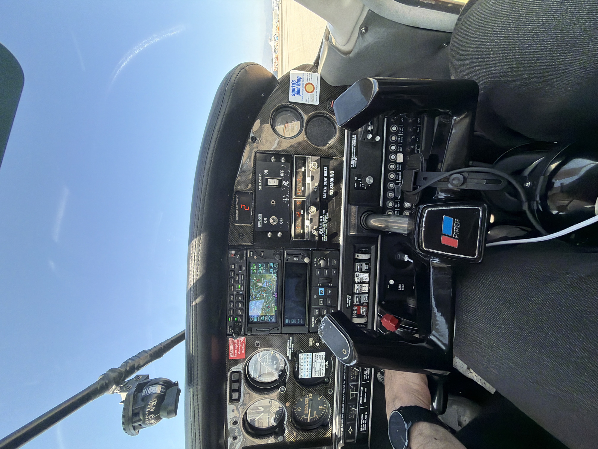 The image shows the cockpit of an aircraft, likely a small plane, with a partial view of the outside sky. The instrument panel is the main focus, featuring various gauges, switches, and avionics equipment. A compass is mounted on the window frame. A man's hand, wearing a watch, is visible near the control stick. A packet of "Sporty's Pilot Shop" carbon monoxide detectors is stuck on the instrument panel. The aircraft is potentially located in Los Angeles, United States of America, but this is inferred from the mention of the city name in the question. The image shows no identifiable landmarks or other features that would help confirm the exact location of the plane in Los Angeles, and it would be difficult to determine based on just the cockpit of the aircraft and a view of the sky.