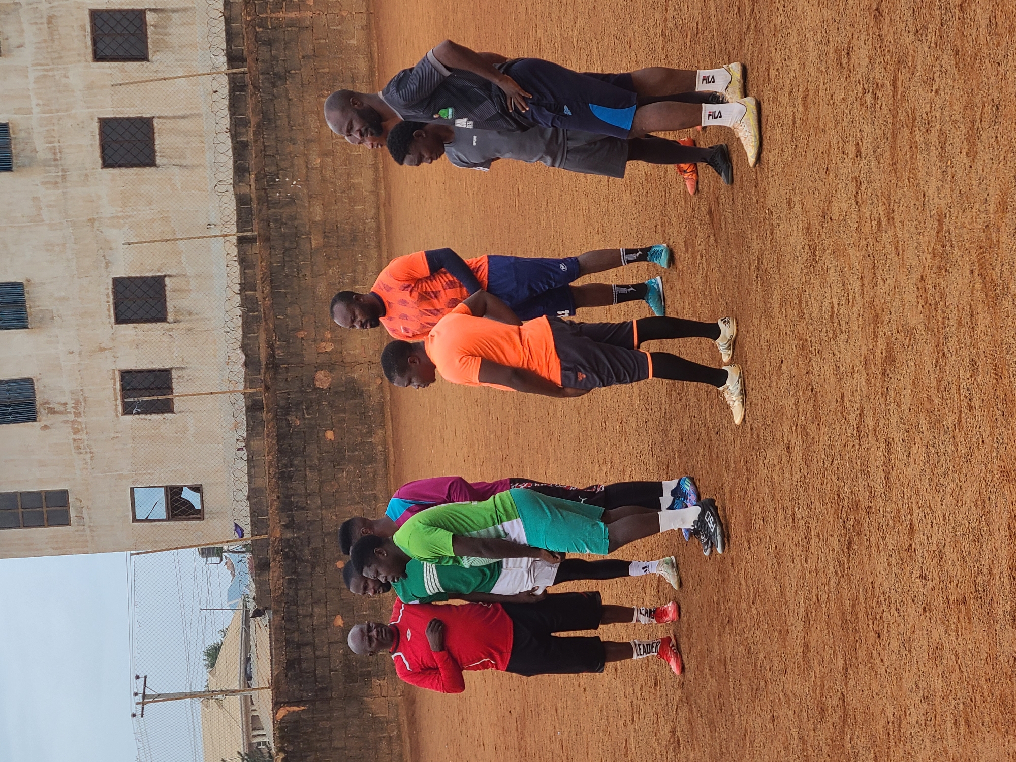 A horizontal medium shot captures a group of six individuals aligned in a straight row on a clay surface. Behind them is a building with a chain-link fence topped with barbed wire. The location appears to be in the city of Unknown, Unknown.
The first person in the line is wearing a red shirt and black shorts. The second is clad in a green shirt and white shorts. The third wears a green shirt with green shorts. The fourth wears a purple shirt and black shorts. The fifth individual is wearing an orange shirt and black shorts. The sixth person is in an orange shirt and blue shorts.
Further back, two more individuals are visible. The first is wearing a gray shirt and dark shorts. The second is wearing a gray shirt and gray shorts. They appear to be stretching forward, with their arms hanging straight down. Their heads are slightly bowed.
The environment appears to be a training ground or field on a clear day. A building is behind them, as well as a chain-link fence topped with barbed wire, adding context to the scene.