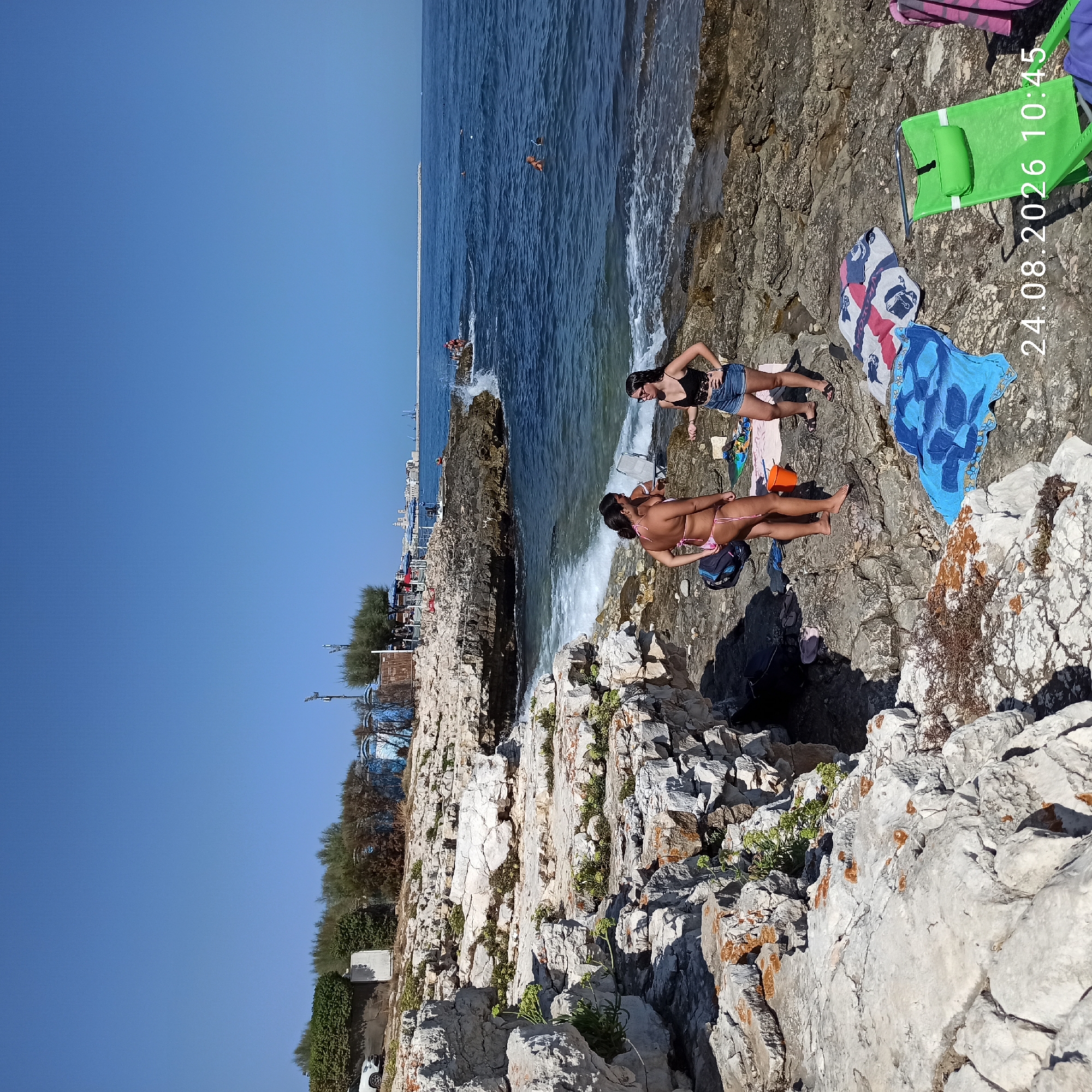The image depicts a rocky coastline in Molfetta, Italy, under a clear blue sky. Three young women are present. One woman, wearing a pink bikini, stands with her back to the camera, facing a second woman who is wearing a black tank top and denim shorts. This second woman is facing slightly away from the camera and appears to be talking to the first woman. A third woman, also in a bikini, stands slightly behind the first woman and to her right, facing the sea.
Several beach towels are spread out on the rocks, and a green beach chair is positioned near the towels. The sea is a deep blue, with waves breaking against the rocks. In the distance, a breakwater extends into the sea, and the cityscape of Molfetta can be seen along the coast, featuring buildings and what appear to be industrial cranes or port structures. Several other individuals are visible in the water, some swimming and one appearing to be snorkeling. The rocky terrain is interspersed with patches of green vegetation. A timestamp "24.08.2026 10:45" is overlaid on the bottom right of the image.