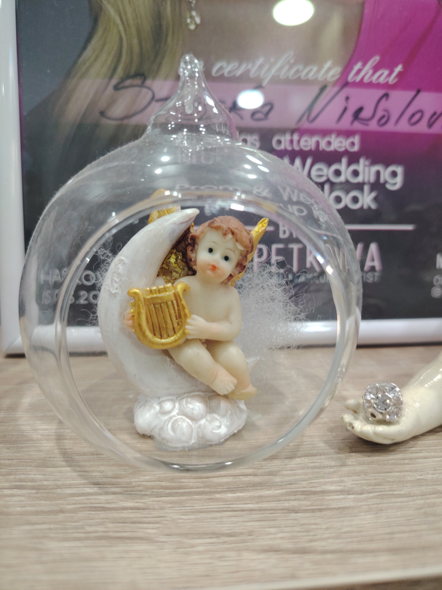 This media file features a close-up shot of a decorative glass ornament containing a small cherub figurine sitting on a crescent moon. The cherub is depicted with golden wings and is holding a golden lyre. The ornament is placed on a wooden surface, likely a table. In the background, a certificate or framed document is visible, with text that appears to relate to a wedding or beauty salon. Some of the visible text includes "certificate that," "has attended," "Wedding Look," and "PETKYA." The overall impression is of a display piece, possibly in a shop or at an event. The lighting suggests an indoor setting, and no specific time of day or weather conditions are discernible. The location context is identified as Haskovo, Bulgaria, which may pertain to the origin or context of the display. There is also a partial view of a decorative hand holding a ring with small rhinestones, adding to the theme of weddings or special occasions.