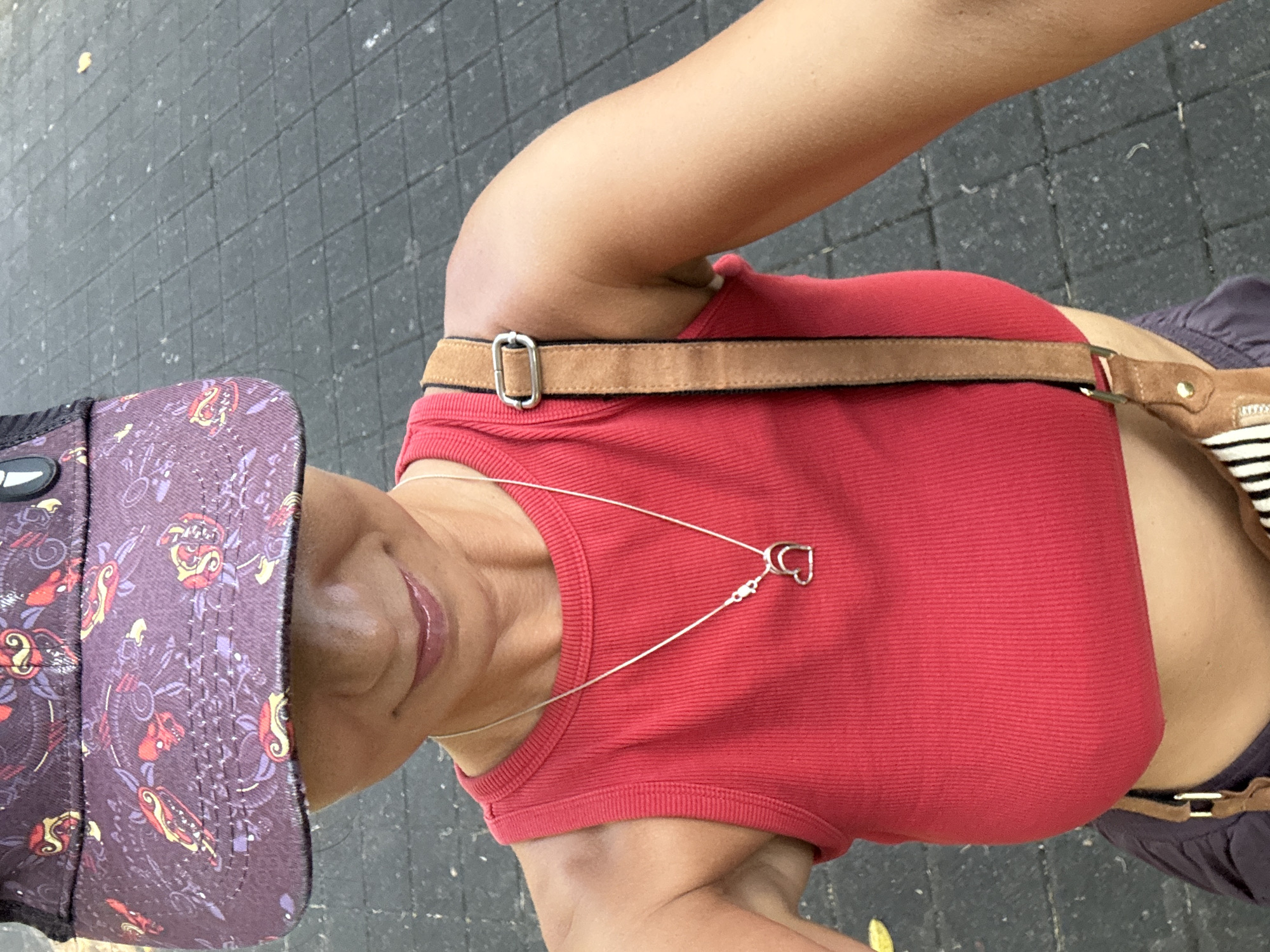 A woman stands on a dark-colored paved surface, potentially a sidewalk or street. She is wearing a red tank top, a brown belt, and a purple patterned cap. A silver heart-shaped pendant hangs from her neck. Part of a bag with black and white stripes is visible on her side. The setting is presumed to be in Kuta Selatan, Indonesia, but there are no visible local landmarks to confirm the location.