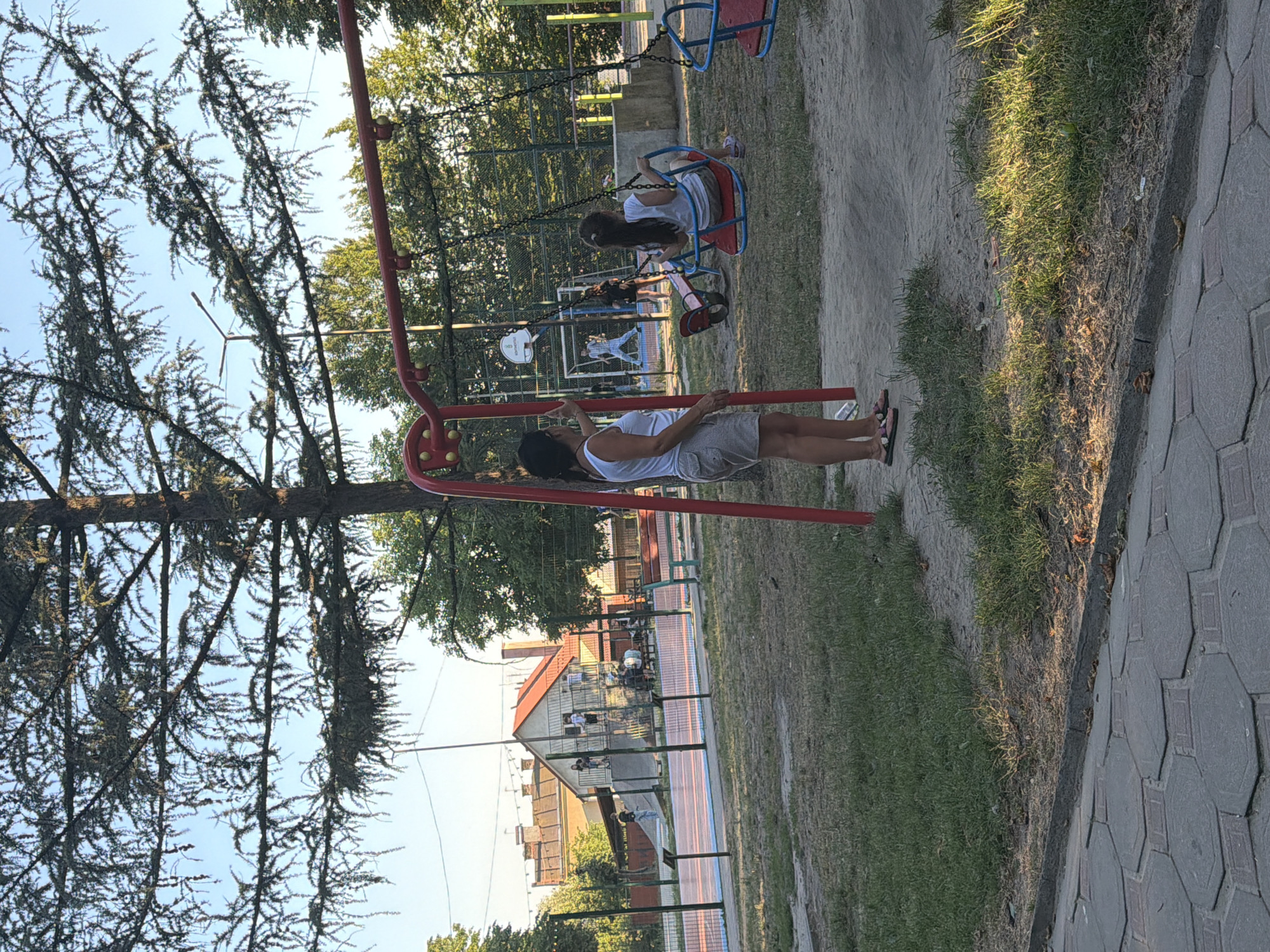 This media file depicts a scene at a playground in Kamianka-Buzka, Ukraine. The primary subjects are people, including at least two children on swings and one adult near a swing set. The setting is an outdoor playground with a red swing set in the foreground. Behind the swing set, there is a fenced-off area that appears to be a sports court, possibly for basketball, with a backboard visible. Further in the background, there are buildings with red roofs and trees.
The main activity is children playing on the swings. The adult in the foreground seems to be interacting with or observing the children. The time of day appears to be late afternoon or early evening, as indicated by the warm lighting and long shadows cast on the ground. The weather seems clear and sunny.
Notable details include the textured pavement on the right side of the image and the sandy surface of the playground. The trees in the foreground are dense and leafy. There is no visible text in the media file. The overall mood is relaxed and casual, typical of a playground setting.