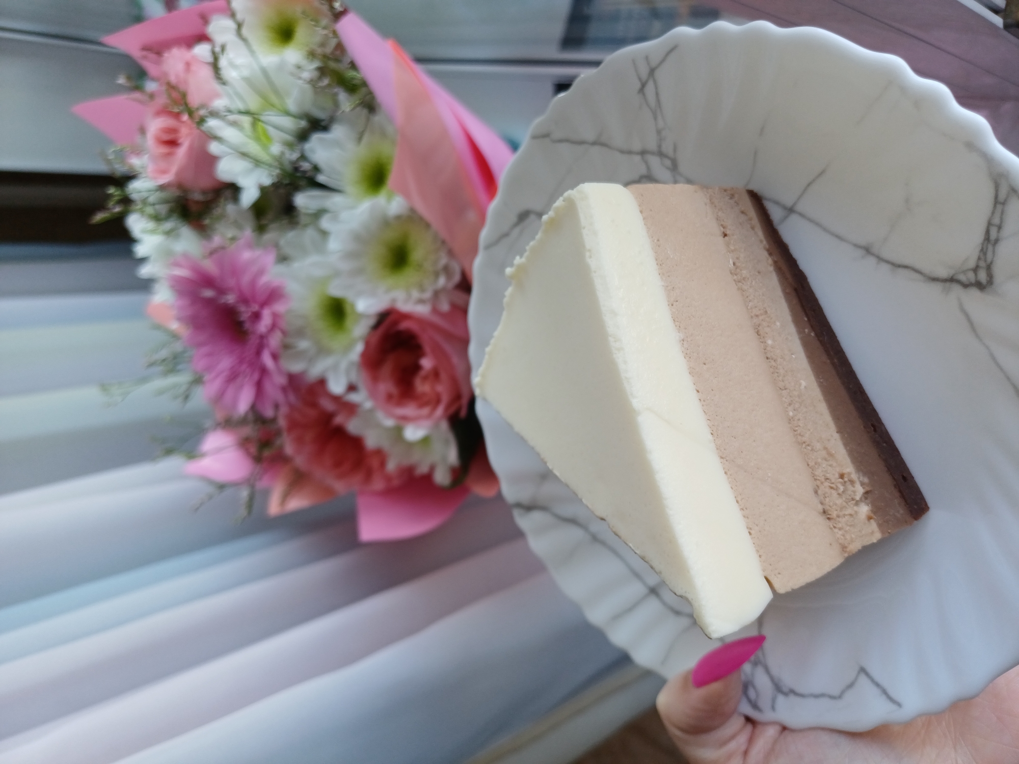 A close-up, top-down shot shows a slice of three-layer cake on a white, marble-patterned plate. The cake has a white top layer, a light brown middle layer, and a dark brown bottom layer, all atop a dark brown base. A hand with bright pink fingernails is holding the plate from the left side of the frame. In the background, out of focus, is a bouquet of flowers wrapped in pink paper. The backdrop consists of white and pale blue vertical blinds. The scene is set indoors.