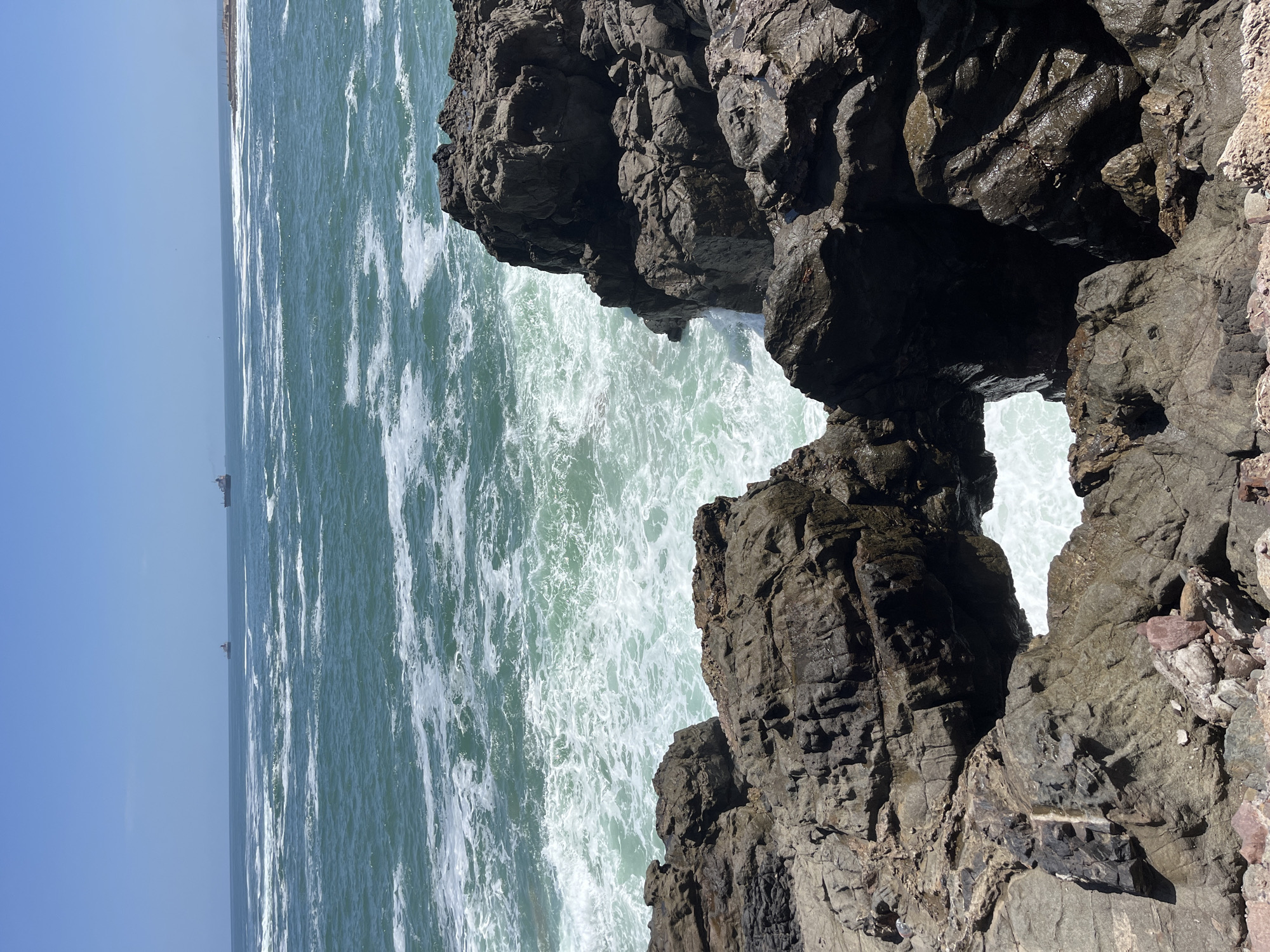 A vertical shot reveals a coastal scene, likely in Arica, Chile, judging by the landscape. Dominating the right side of the image are dark, rocky cliffs, with a natural arch visible through which the turquoise ocean is seen. The sea is turbulent with white foam from breaking waves. In the distance, ships dot the horizon under a clear blue sky. The composition showcases the rugged coastline, typical of the region, and the dynamic interaction between land and sea.