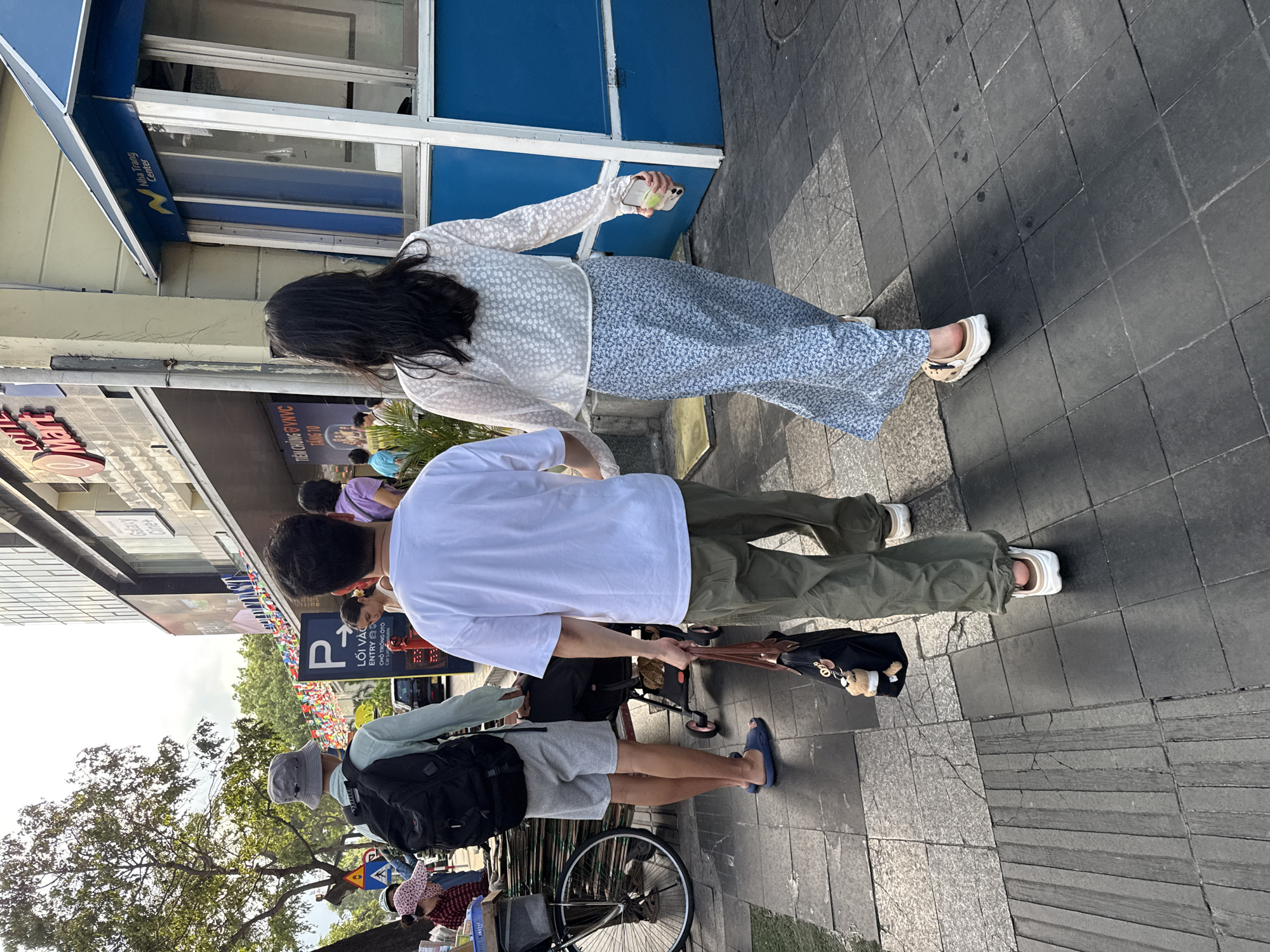 The image shows a street scene in Nha Trang Ward, Vietnam, with a focus on two individuals walking away from the camera. A man in a light blue long-sleeved shirt, grey shorts, and a grey bucket hat is walking on the left, carrying a black backpack. He is wearing blue flip-flops. To his right, a woman with long dark hair is wearing a white sheer floral-patterned top and a long blue floral-patterned skirt. She is wearing white slip-on sandals and holding a smartphone in her right hand. The man is pulling a black stroller. A black tote bag with a teddy bear charm is hanging from the man's right hand. In the background, a building with a "LOTTE Mart" sign is visible, along with a blue and white kiosk labeled "Nha Trang Center." Several people and a bicycle are also present in the background. The scene takes place on a paved sidewalk with visible cracks and patterns.