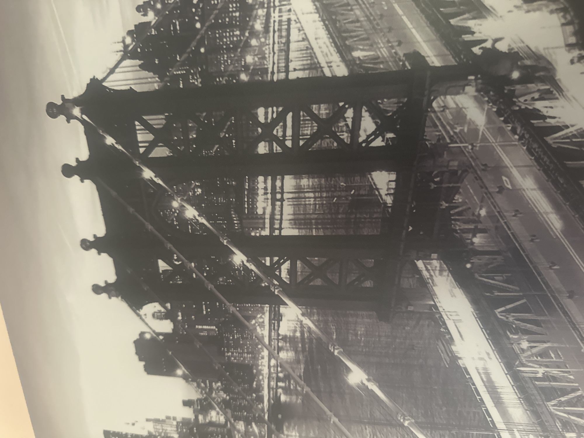 The image is a black and white photograph of the Manhattan Bridge in New York City, viewed from a low angle looking up at the bridge's support structure. The bridge's latticework, cables, and roadways are prominent, along with city lights in the background. The photo captures the industrial and architectural detail of the bridge at night.