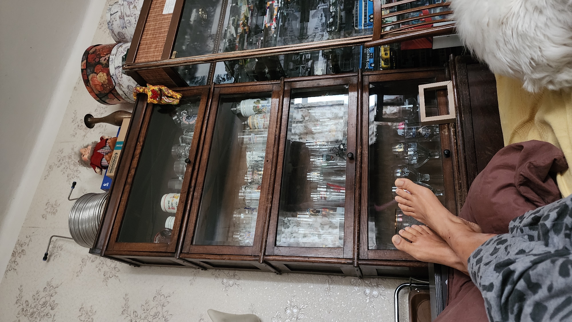 The image depicts an indoor scene, likely a living room, featuring a tall wooden display cabinet filled with various glassware, predominantly beer glasses. The cabinet is multi-tiered, with glass doors on each level. On top of the cabinet, there are decorative items including a doll, a vase, and floral hatboxes. To the right of the cabinet, another display case is visible, filled with model vehicles. In the foreground, a person's bare feet are resting on a pillow, suggesting they are lying down. The wall behind the cabinet is covered in floral patterned wallpaper. The scene appears to be a personal collection displayed in a home. There is no indication of the location being in Moscow, Russia.