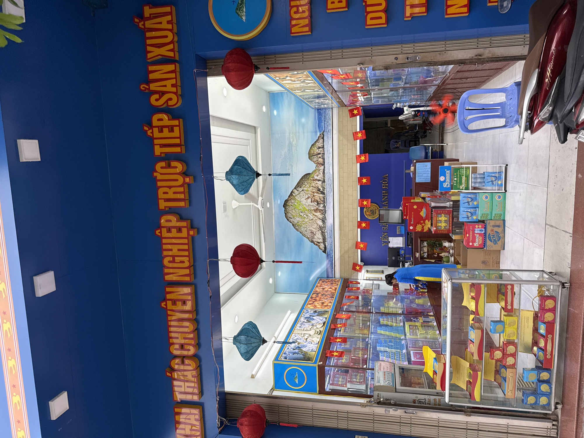 The image shows the exterior of a shop in Nha Trang, Vietnam. The shop has a bright blue facade with a large yellow and red sign at the top that reads "KHAI THÁC CHUYÊN NGHIỆP TRỰC TIẾP SẢN XUẤT". Below the sign, the shop entrance is visible, with red and blue lanterns hanging from the ceiling. Inside, shelves are stocked with various products, including what appear to be food items and drinks. A woman in a blue and yellow outfit is visible working inside the shop. A glass display case in front of the shop contains boxes of products. A red motorcycle is parked in the foreground. The overall impression is of a busy retail establishment in a Vietnamese city.